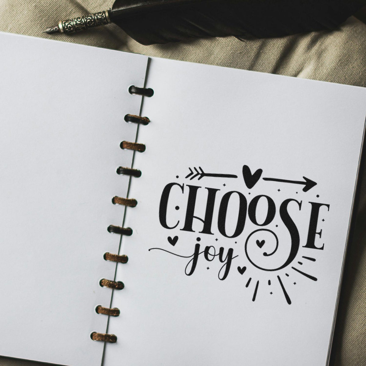 Black CHOOSE joy stamp design on a white spiral notebook, with hearts, arrows, and decorative seal-style lettering.