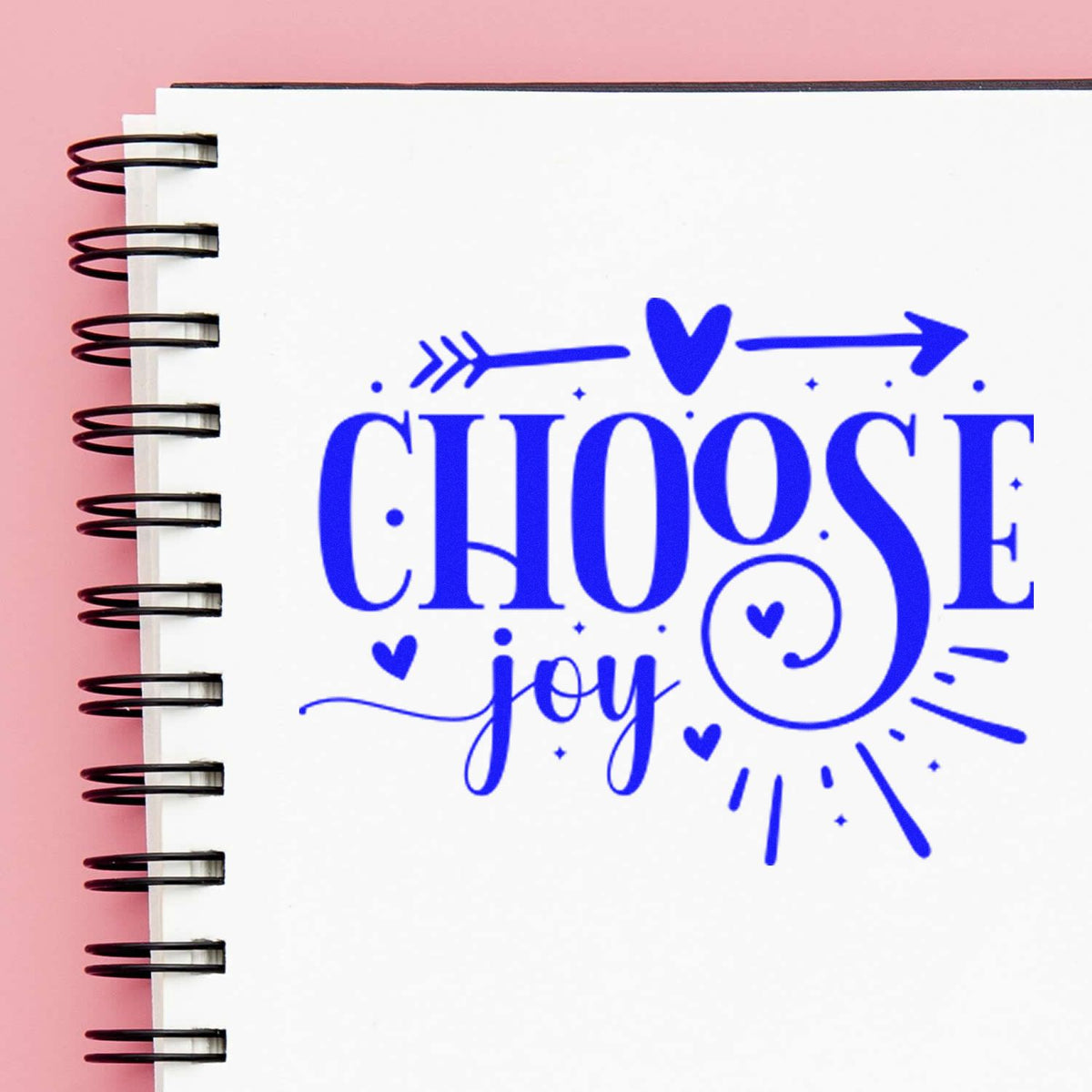 Blue stamp seal reading CHOOSE joy with hearts and arrows on a white spiral notebook, pink background.