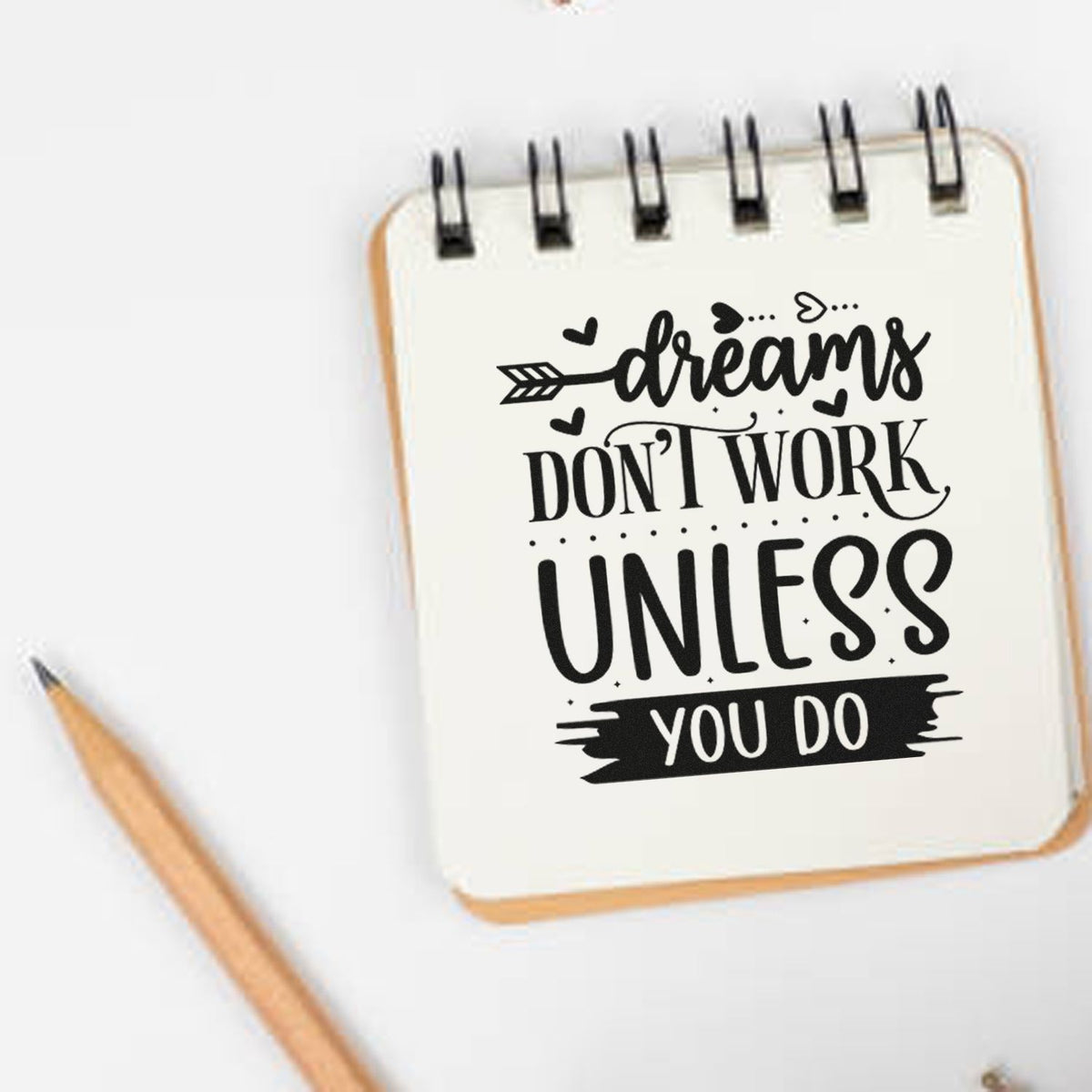 Black stamp-style quote on cream spiral notebook with pencil: Dreams dont work unless you do.