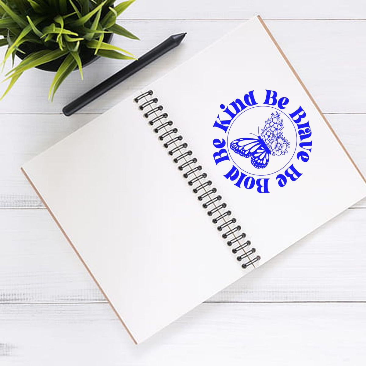 Blue custom stamp seal with butterfly and floral design on a white spiral notebook, beside a black pencil and green plant.