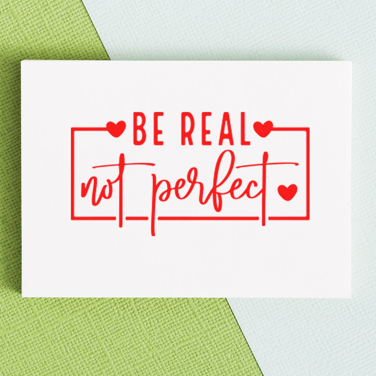 Red stamp seal reading Be real not perfect with hearts on a white card, set on a green and white textured background.
