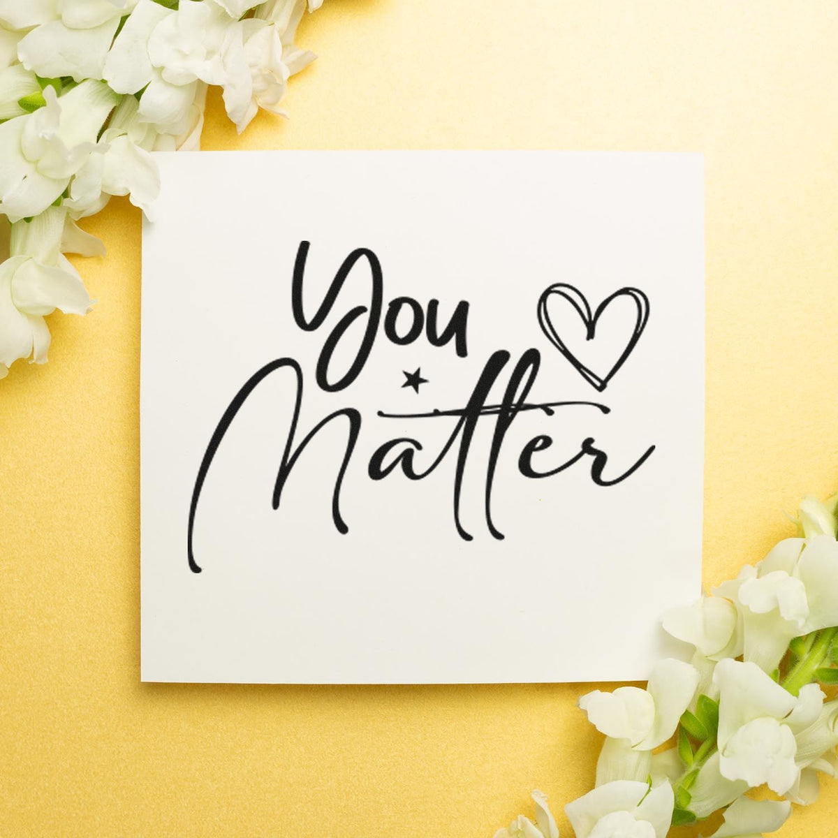 Black You Matter stamp and seal design with heart on white card, framed by white flowers on a soft gold background.