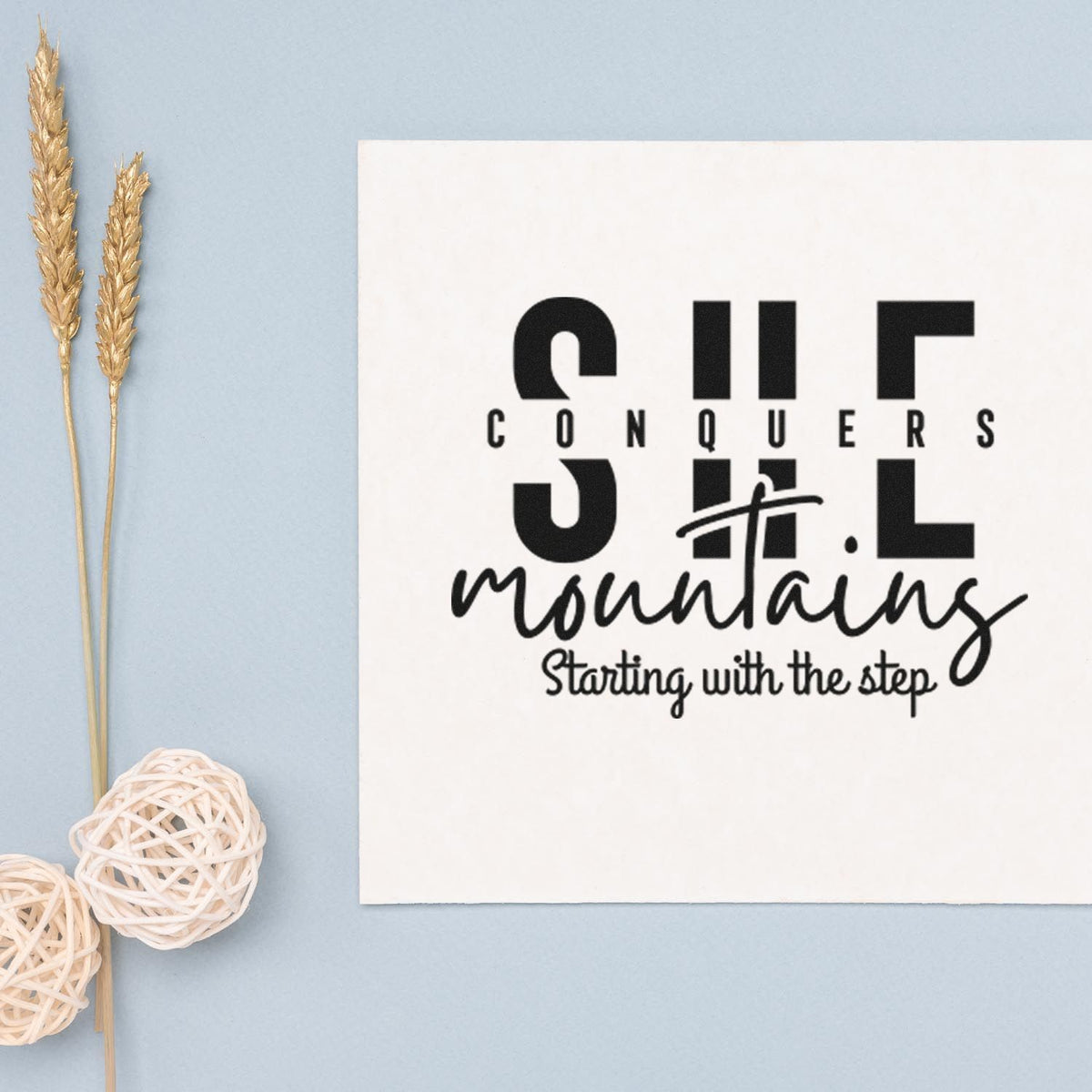 Black custom stamp and seal design on white card reading Conquers Mountains, on a pale blue background with gold wheat accents.