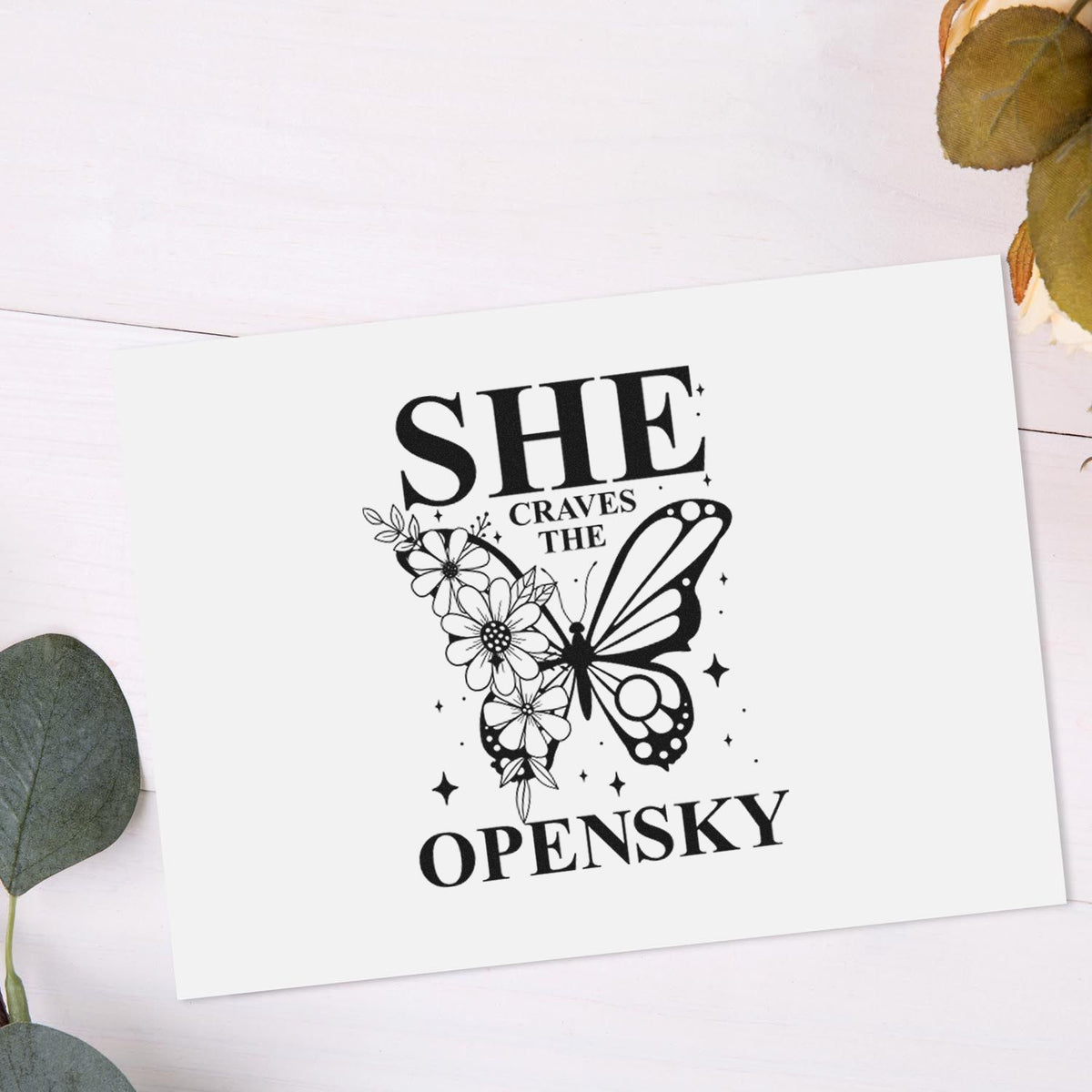Black custom stamp seal with butterfly and floral design, reading SHE CRAVES THE OPEN SKY, on white card with eucalyptus leaves.