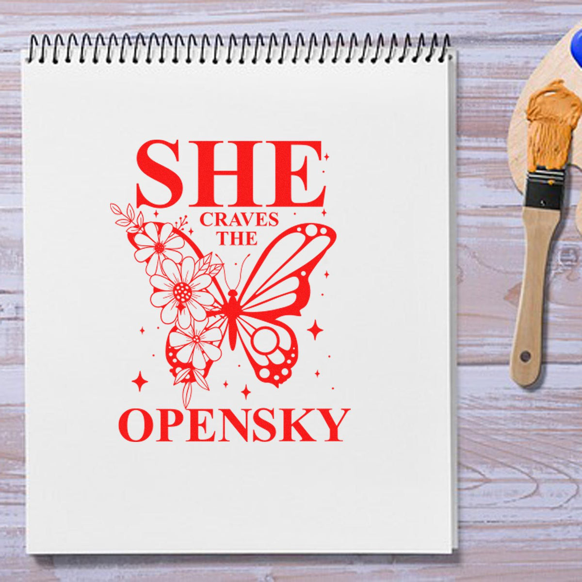 Red butterfly floral stamp design reading SHE CRAVES THE OPENSKY on a white spiral notebook, with a seal brush at right.