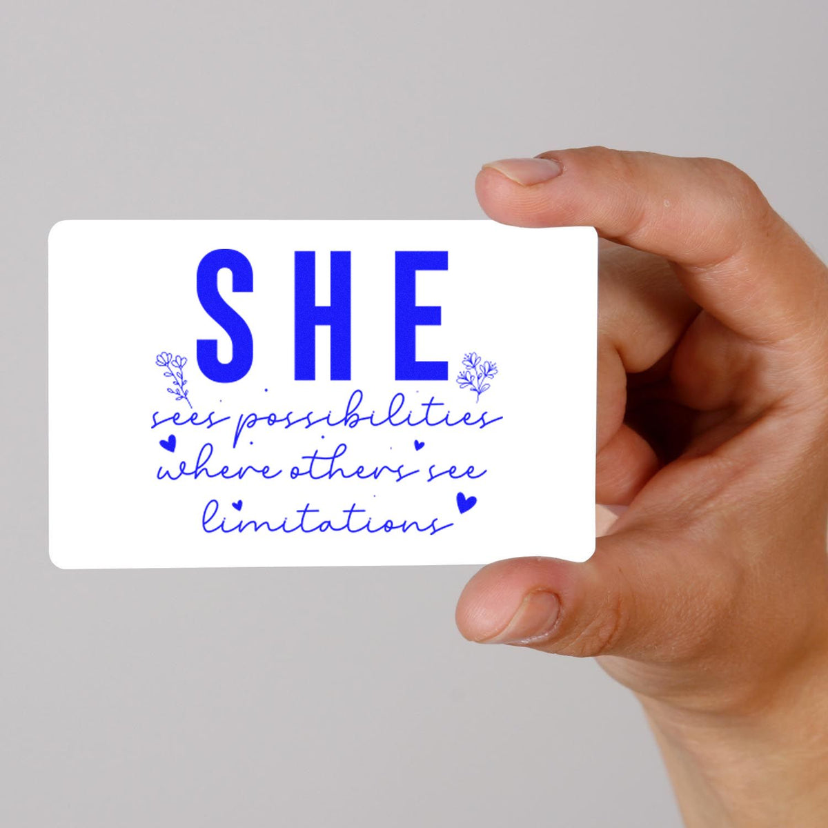 Hand holding white stamp and seal card with blue SHE quote and floral accents: sees possibilities where others see limitations.