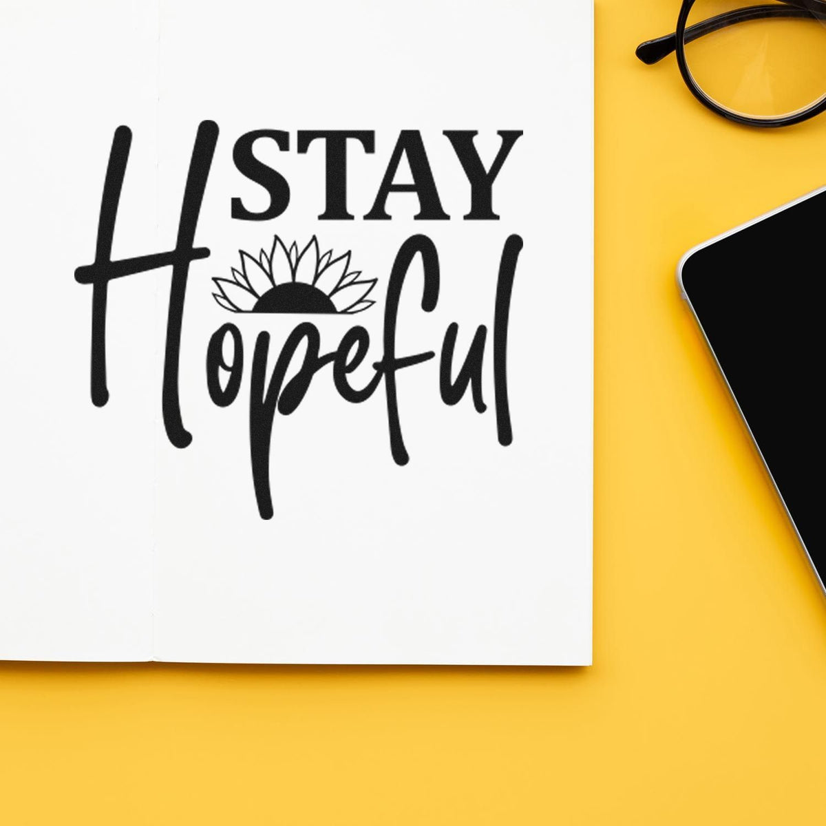Black Stay Hopeful stamp and seal design with sunflower on white paper, set on a yellow background beside glasses and a phone.
