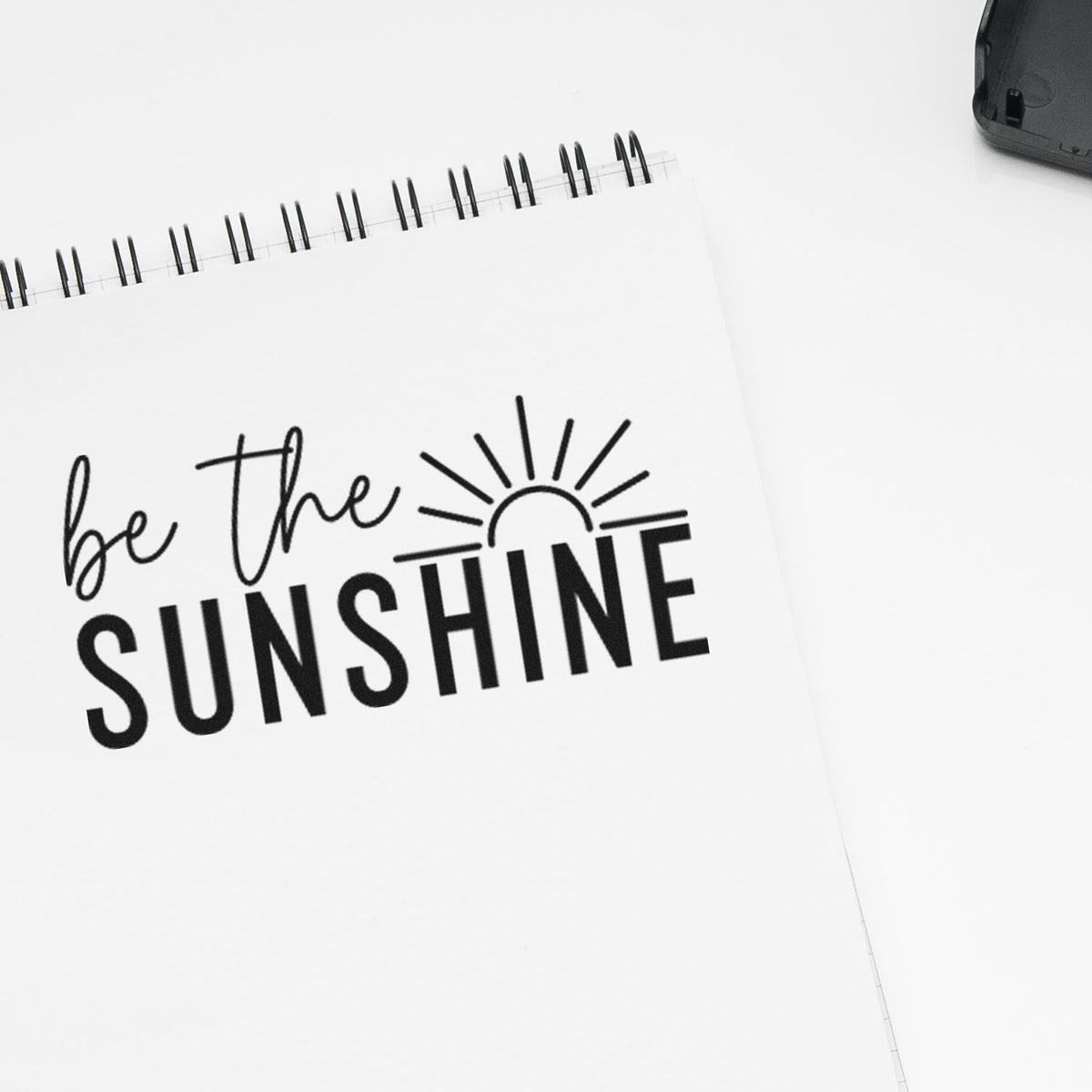 Black custom stamp and seal design on white notebook page reading be the SUNSHINE with sun icon.