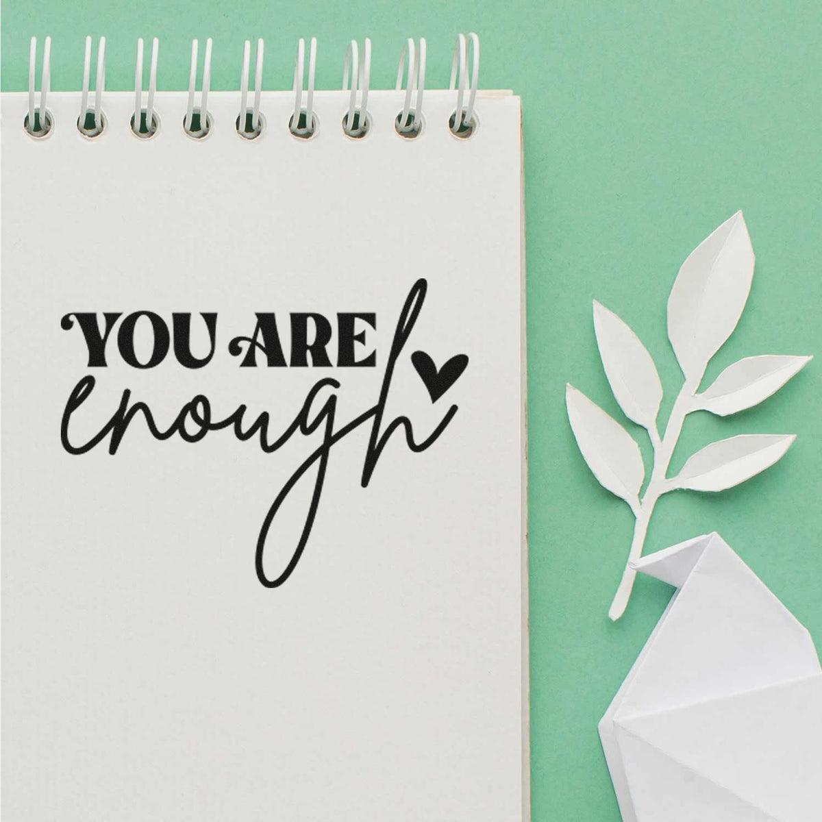 Black You are enough stamp on white spiral notebook, with white paper leaf seal and folded geometric seal on mint green.
