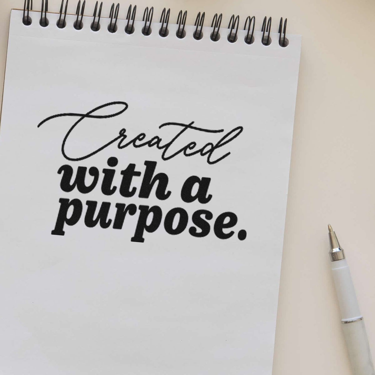Black custom stamp reading Created with a purpose on a white spiral notebook, with a white pen on a beige background.