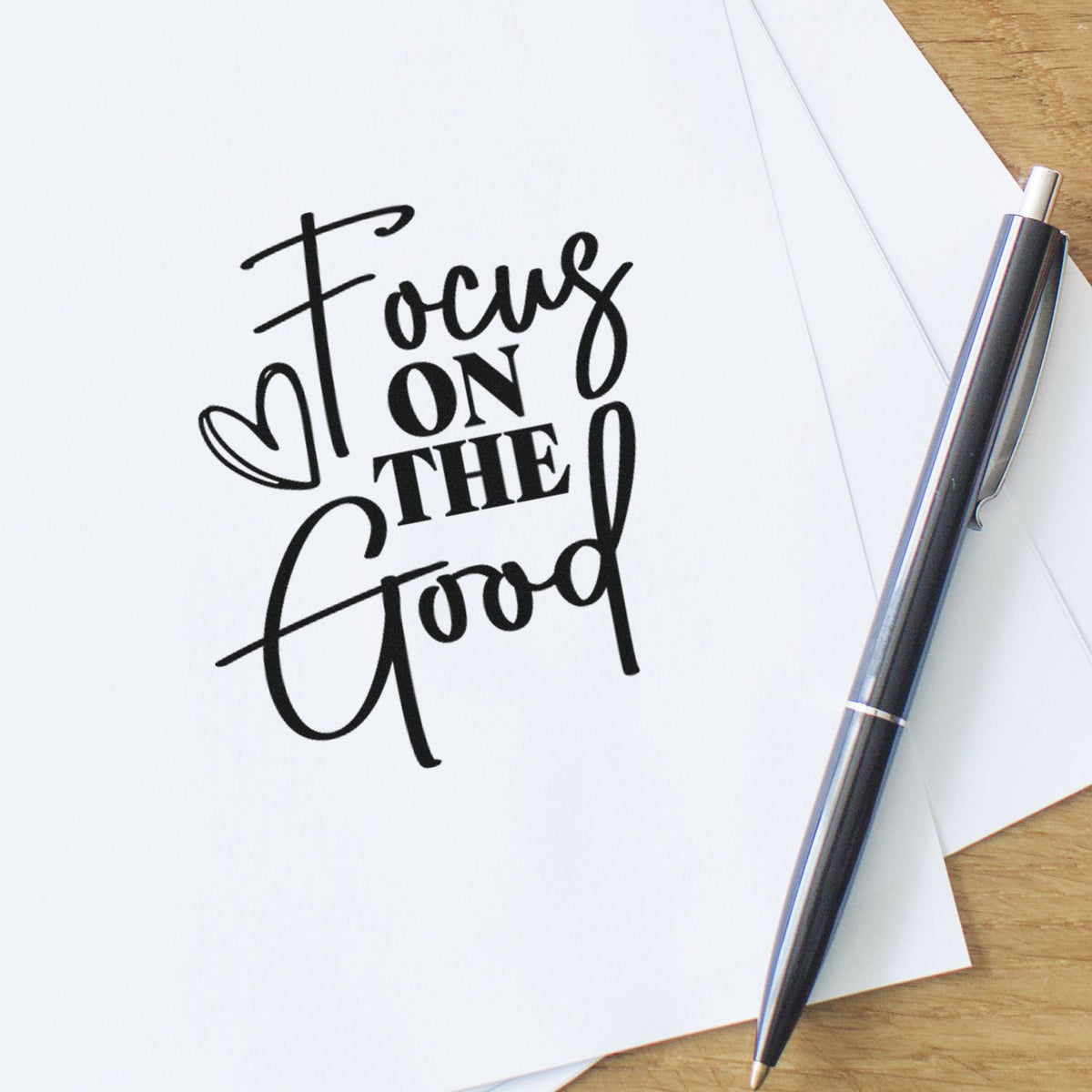 Black custom stamp and seal reading Focus on the Good on white paper, with a sleek black pen on a wood desk.