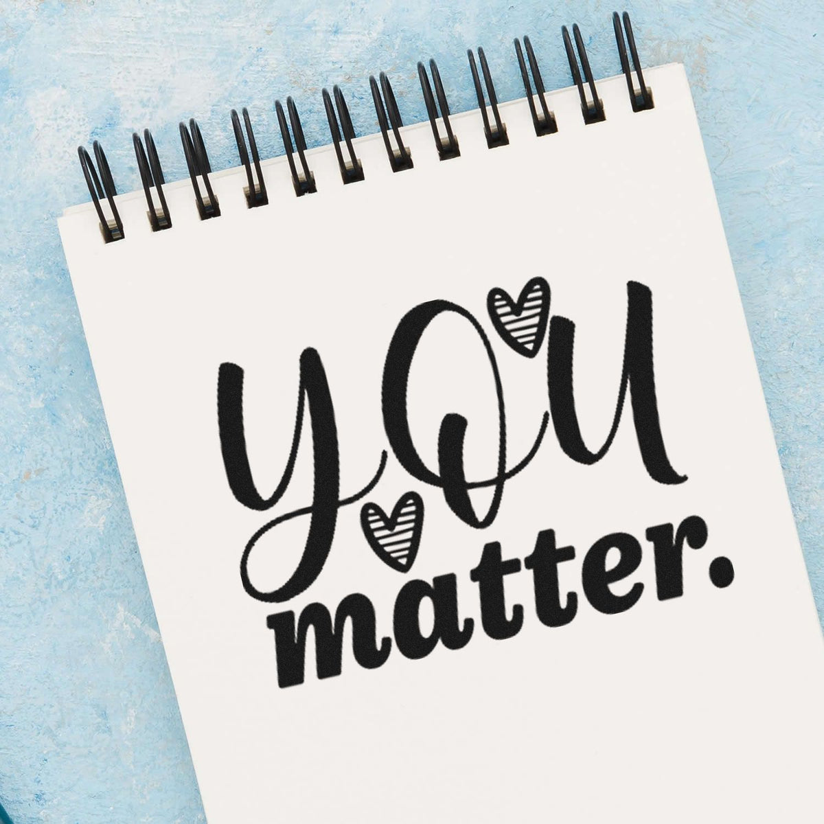 Black you matter motivational stamp design on a white spiral notepad with heart accents, on a light blue textured background.