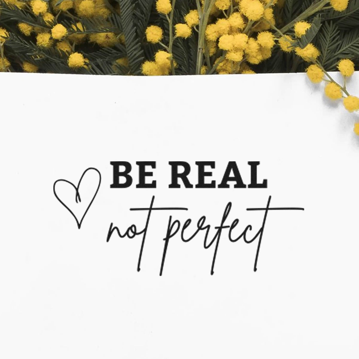 Black stamp and seal design reading Be real not perfect with heart outline, on white card and yellow flowers.