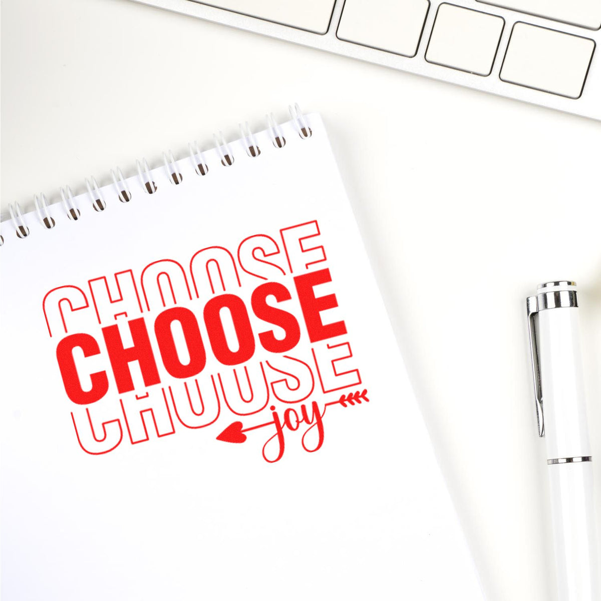 Red custom stamp/seal reading "Choose Choose Joy" on white spiral notebook beside silver pen and keyboard.