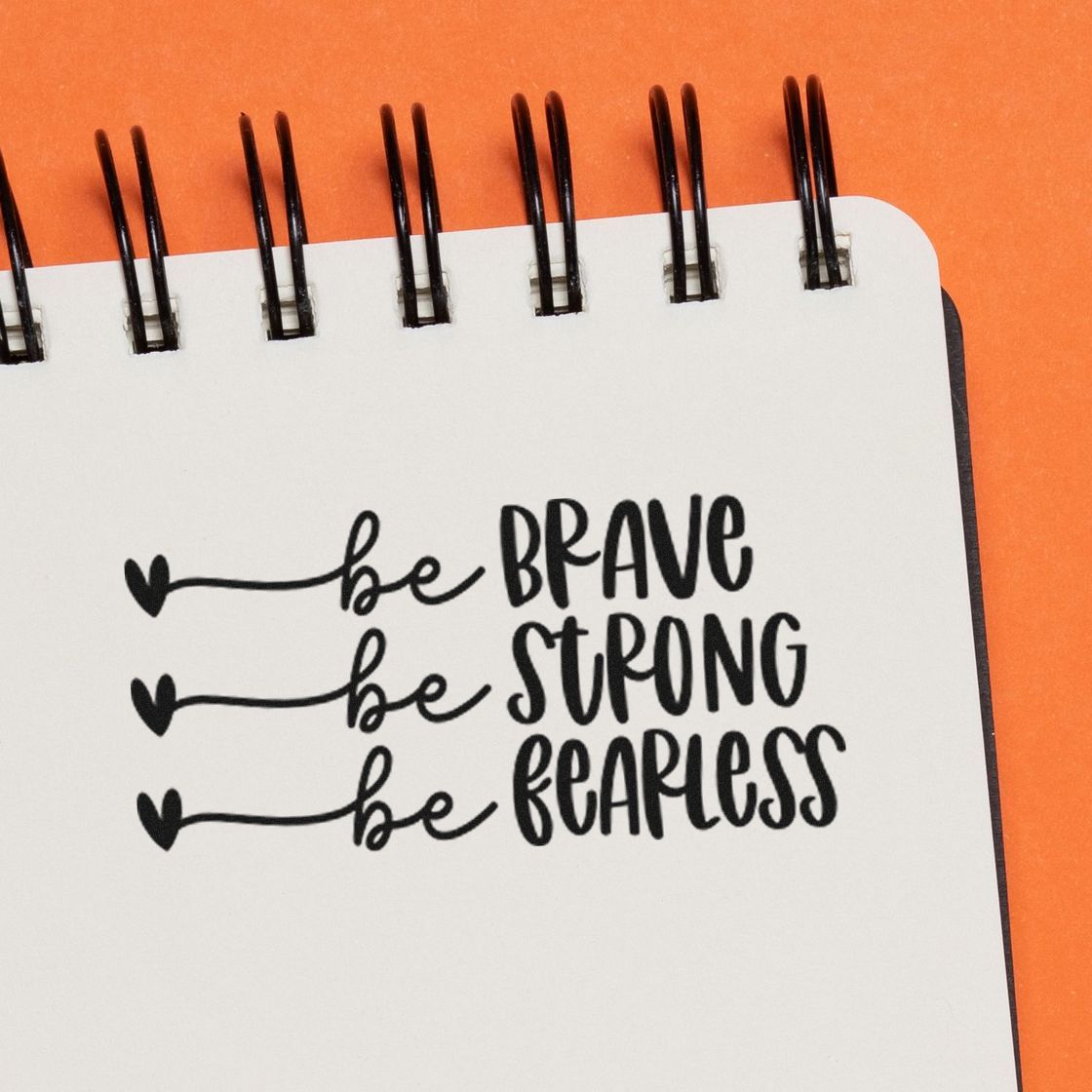 Black stamp-style motivational lettering on a white spiral notebook: be brave, be strong, be fearless, with heart accents on orange background.