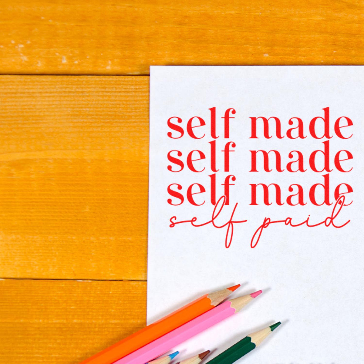 Red self made/self paid stamp design on white paper with colorful pencils on a wooden table.