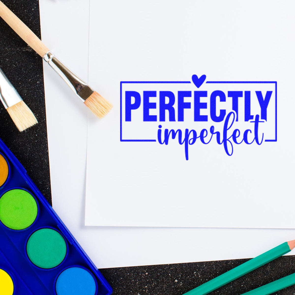 Blue Perfectly imperfect stamp and seal on white paper, with paintbrushes, watercolor palette, and colored pencils.