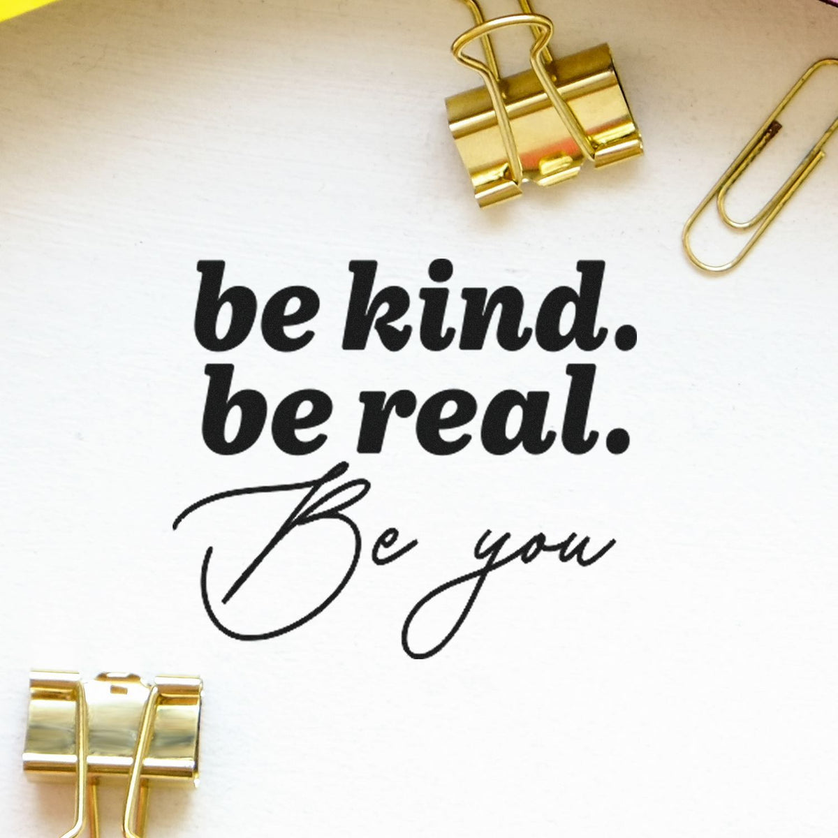 Black motivational stamp and seal design on white paper, framed by gold binder clips, with be kind. be real. Be you.