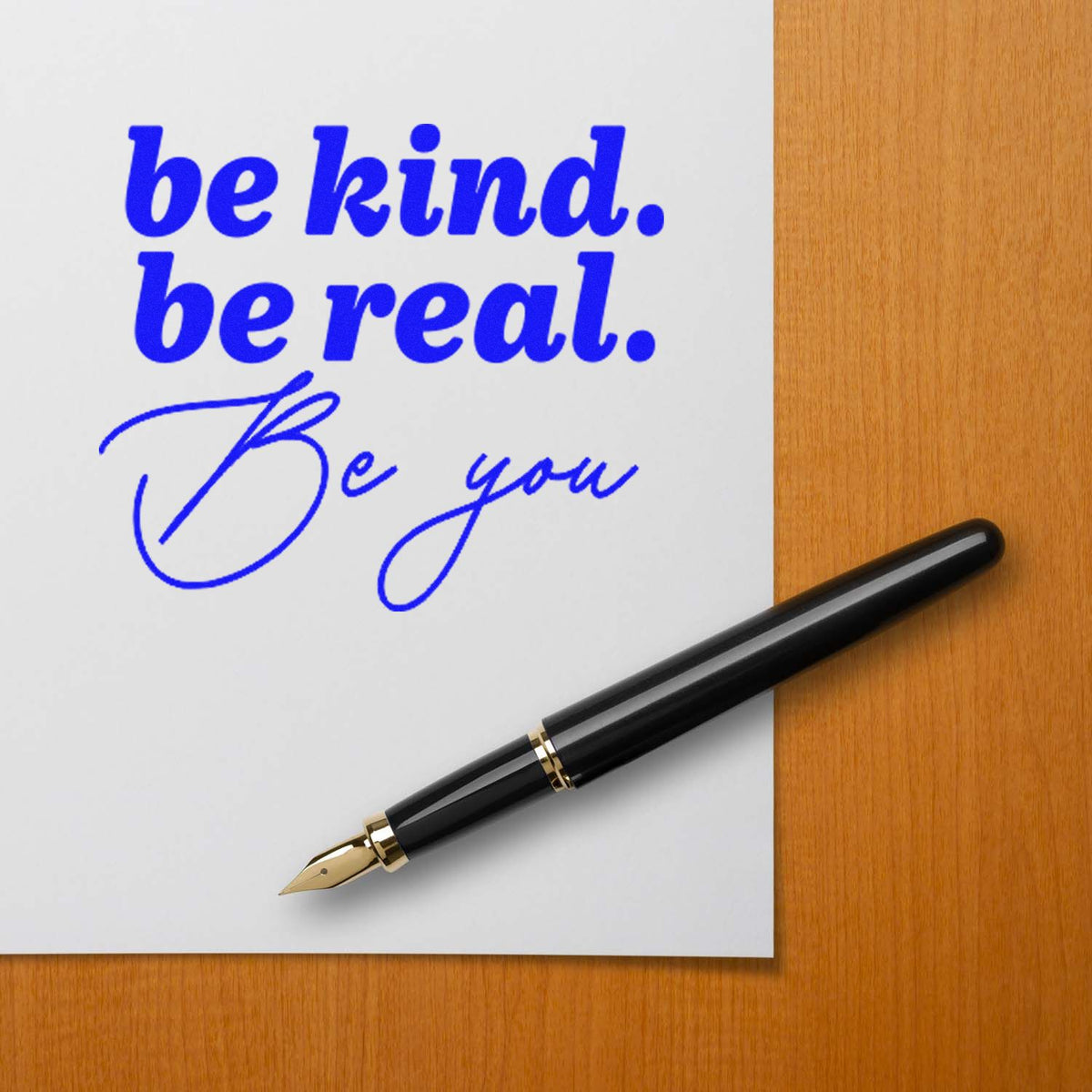 Blue self-inking stamp reading be kind. be real. Be you beside a black fountain pen on a wood desk.