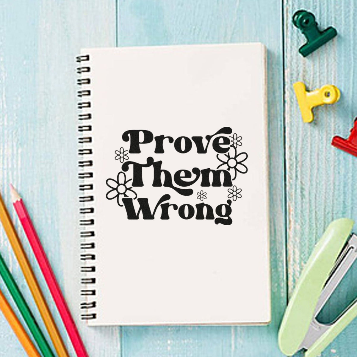White spiral notebook with a black floral stamp reading Prove Them Wrong, plus colorful binder clips and pencils on blue wood.