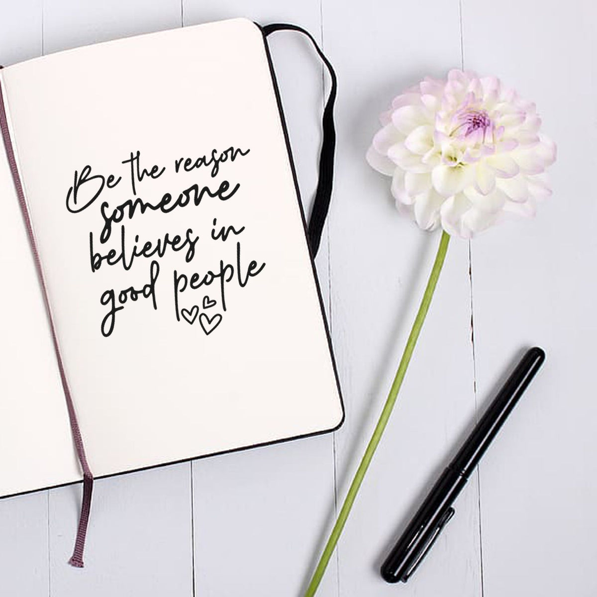 Stamp and seal product flat lay: cream notebook with black handwritten quote, white flower, and black pen on pale wood.