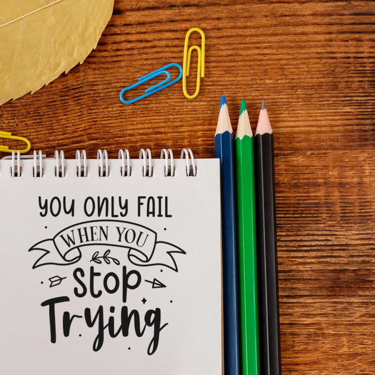 Motivational stamp seal on notebook reading You only fail when you stop trying, beside blue, green and black pencils on a wood desk.