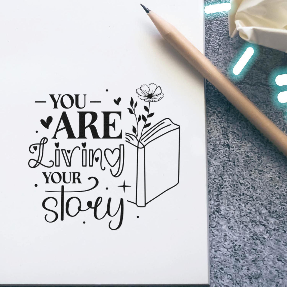 Black stamp-and-seal design on white notebook reading You are living your story, with book, flower, and hearts.
