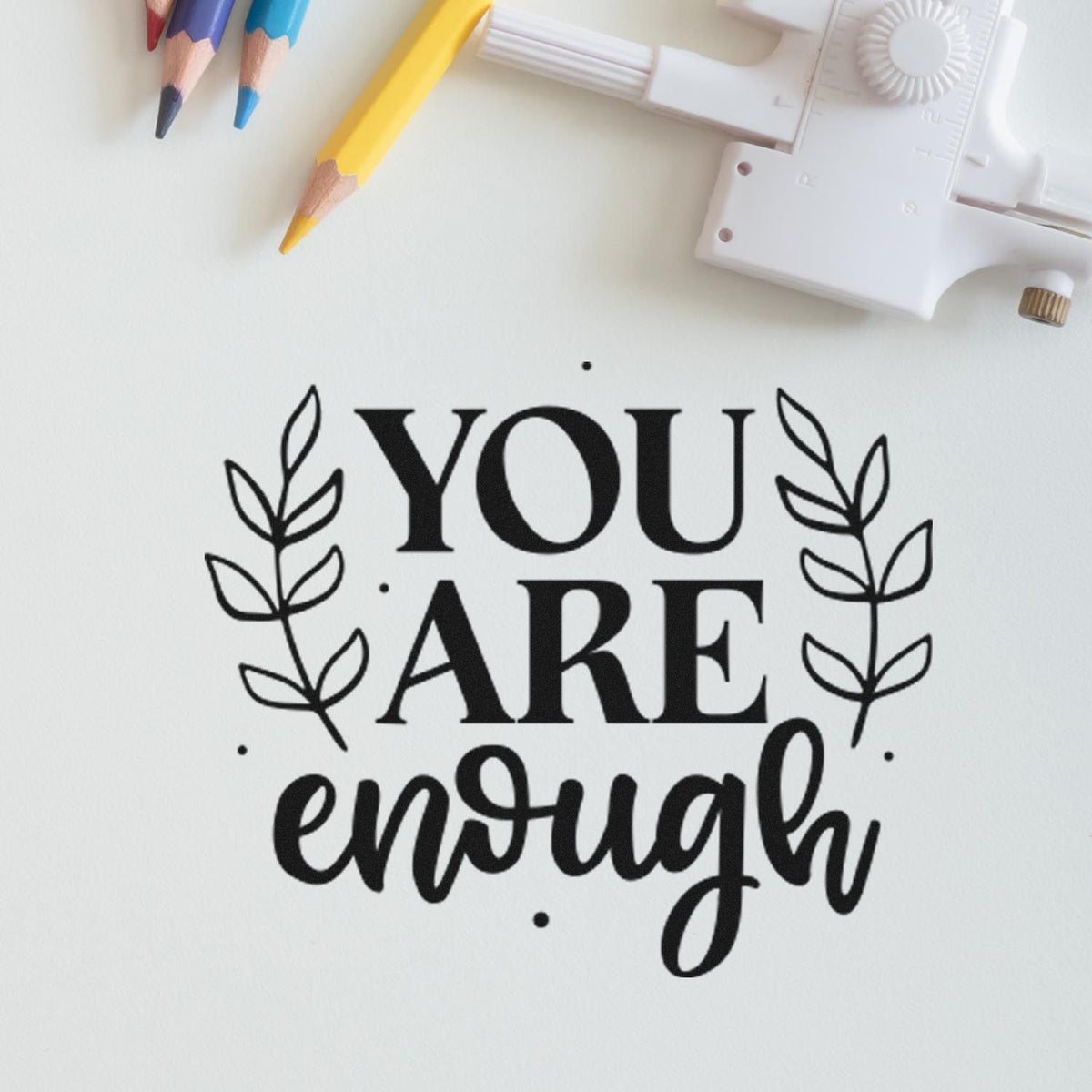 Black stamp reading YOU ARE enough with leaf accents on white paper, beside colored pencils and a white seal tool.