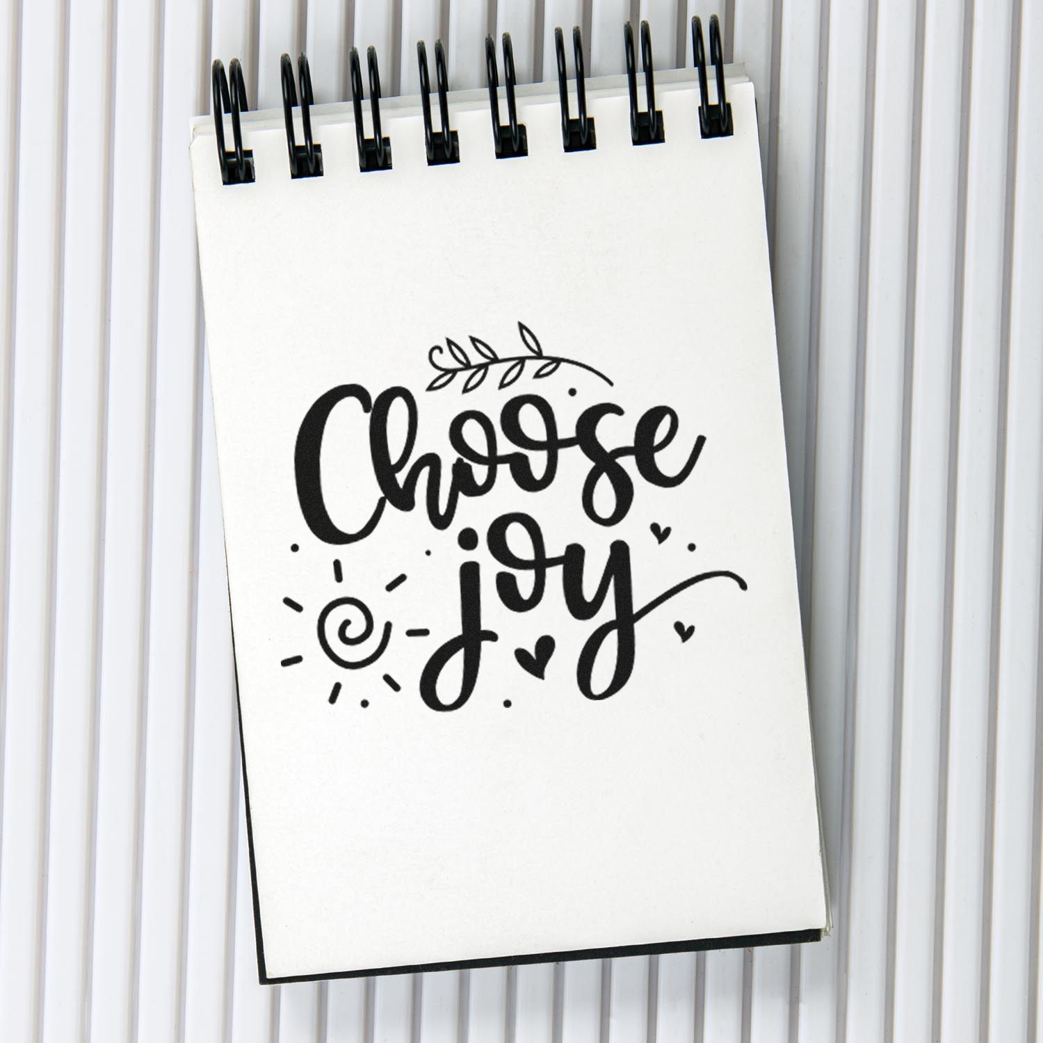 Black Choose Joy stamp and seal design on white spiral notebook, with leaf, heart, and sun doodles.