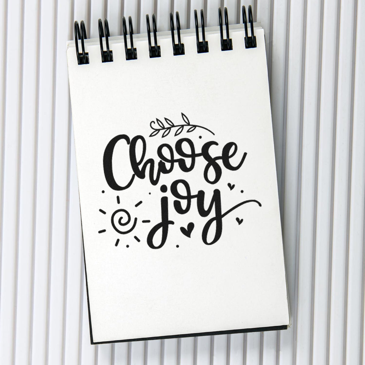 Black Choose Joy stamp seal design with hearts, sun, and leaf on a white spiral notepad.