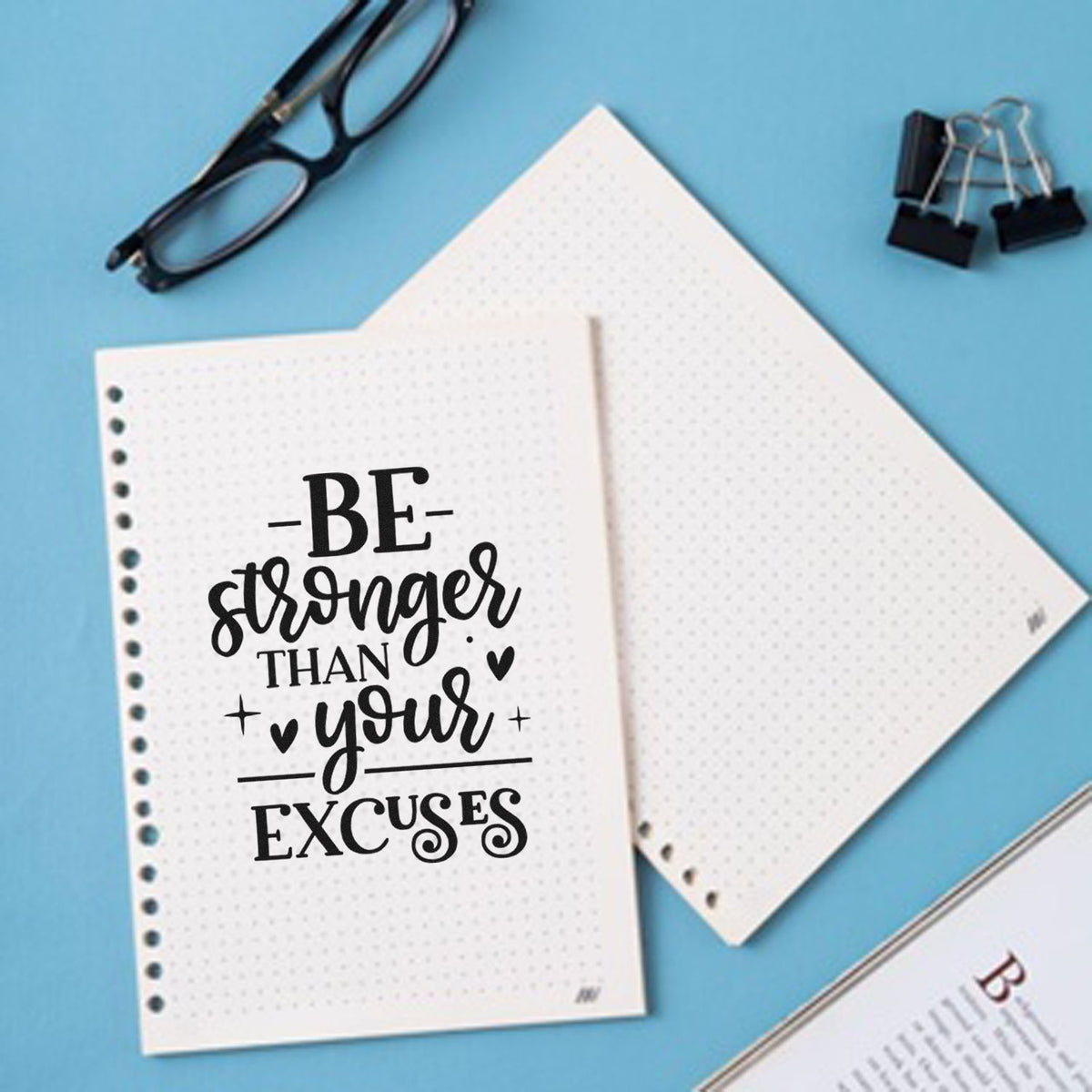 Black stamp-style design reading Be stronger than your excuses on white dotted notebook pages, with glasses and binder clips on blue.