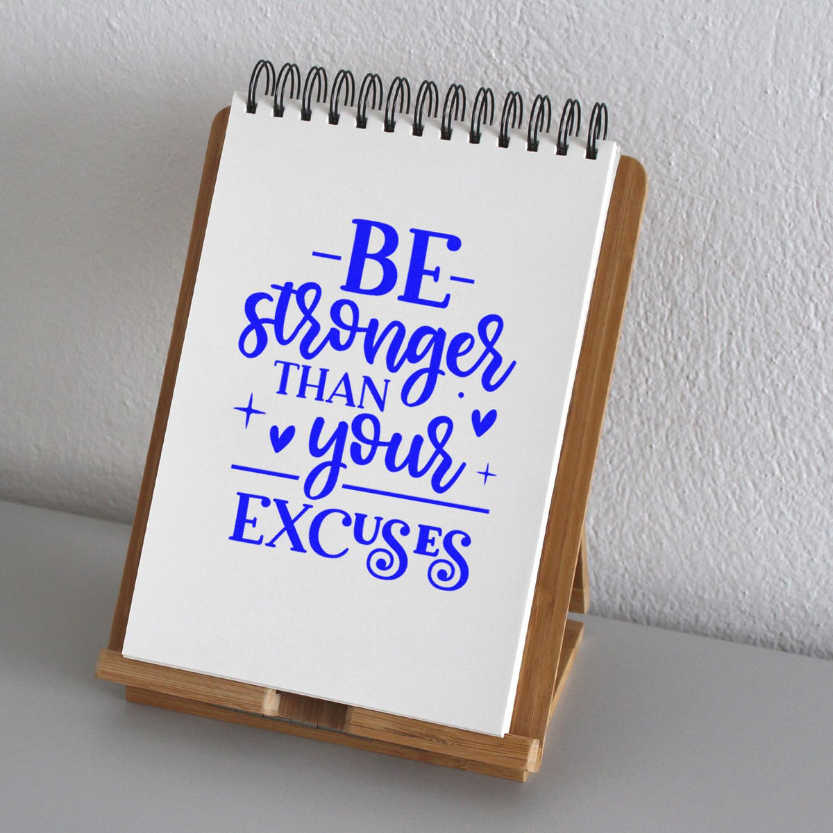 Blue custom stamp design reading Be stronger than your excuses on a white spiral notepad with wooden stand.