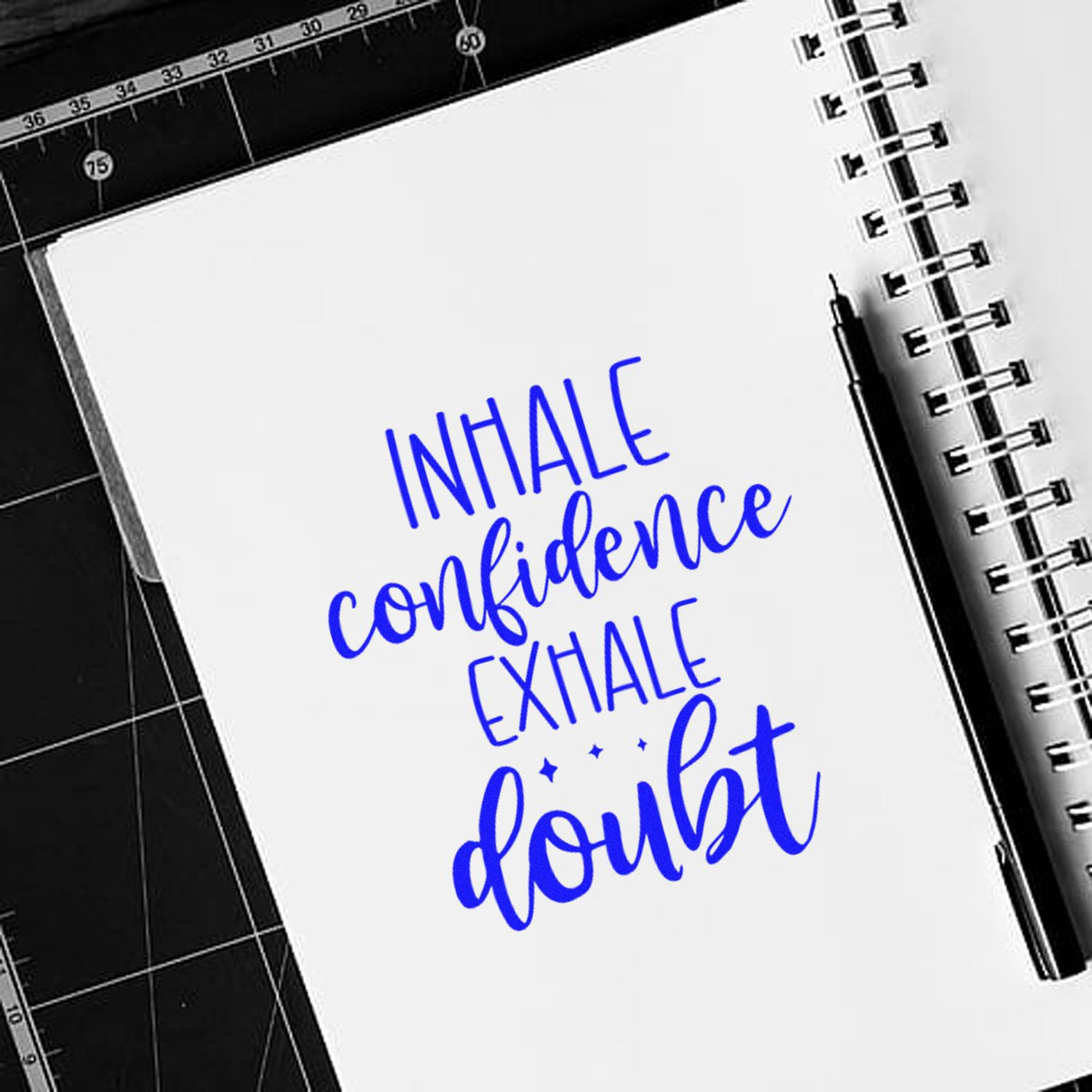 Blue stamp and seal design on a white spiral notebook, reading inhale confidence, exhale doubt, beside a black pen.