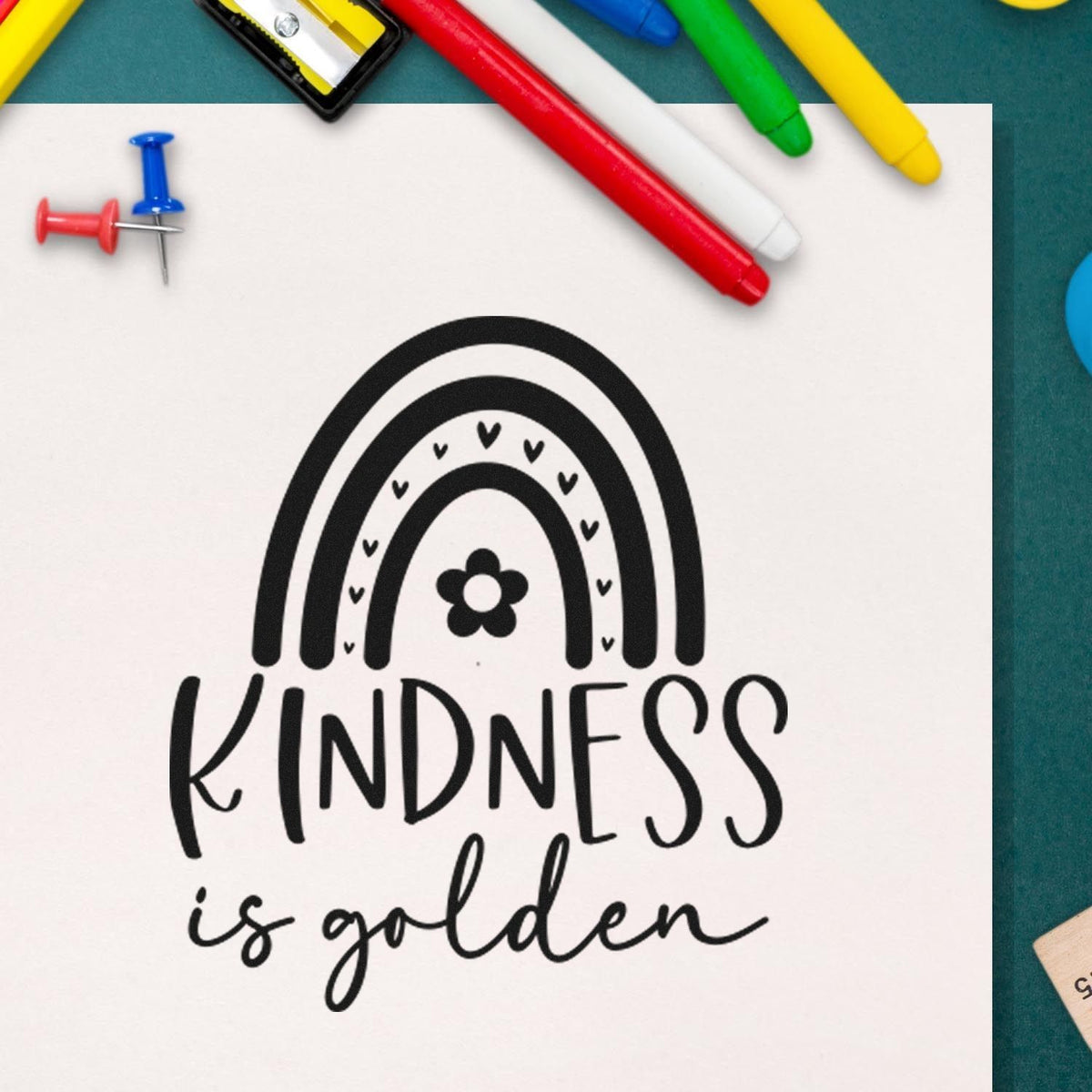 Black kindness stamp and seal design with rainbow, hearts and flower, on white paper with colorful pens and push pins.