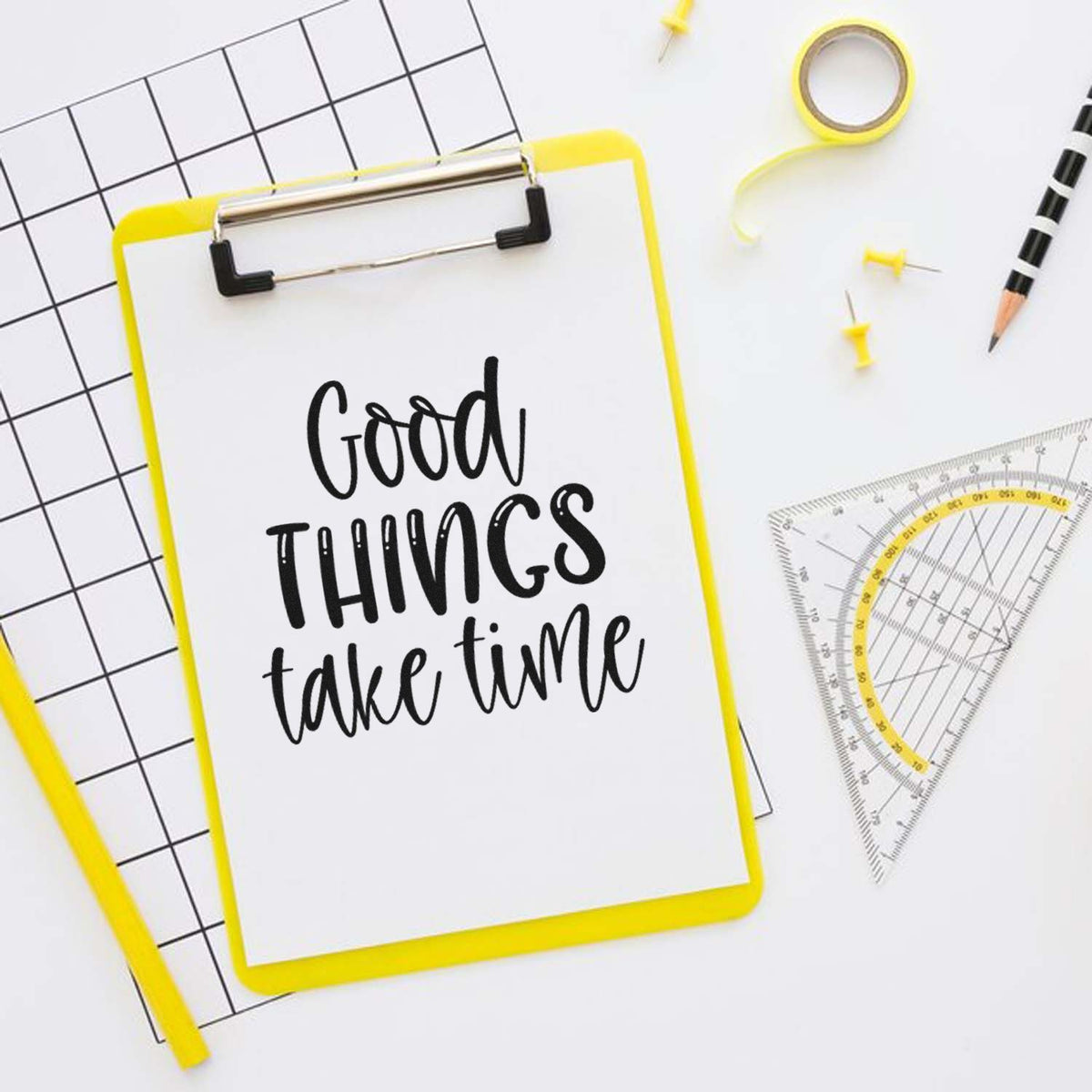 Yellow stamp and seal clipboard with black Good things take time text, on white with grid paper, tape, pins, and drafting tools.