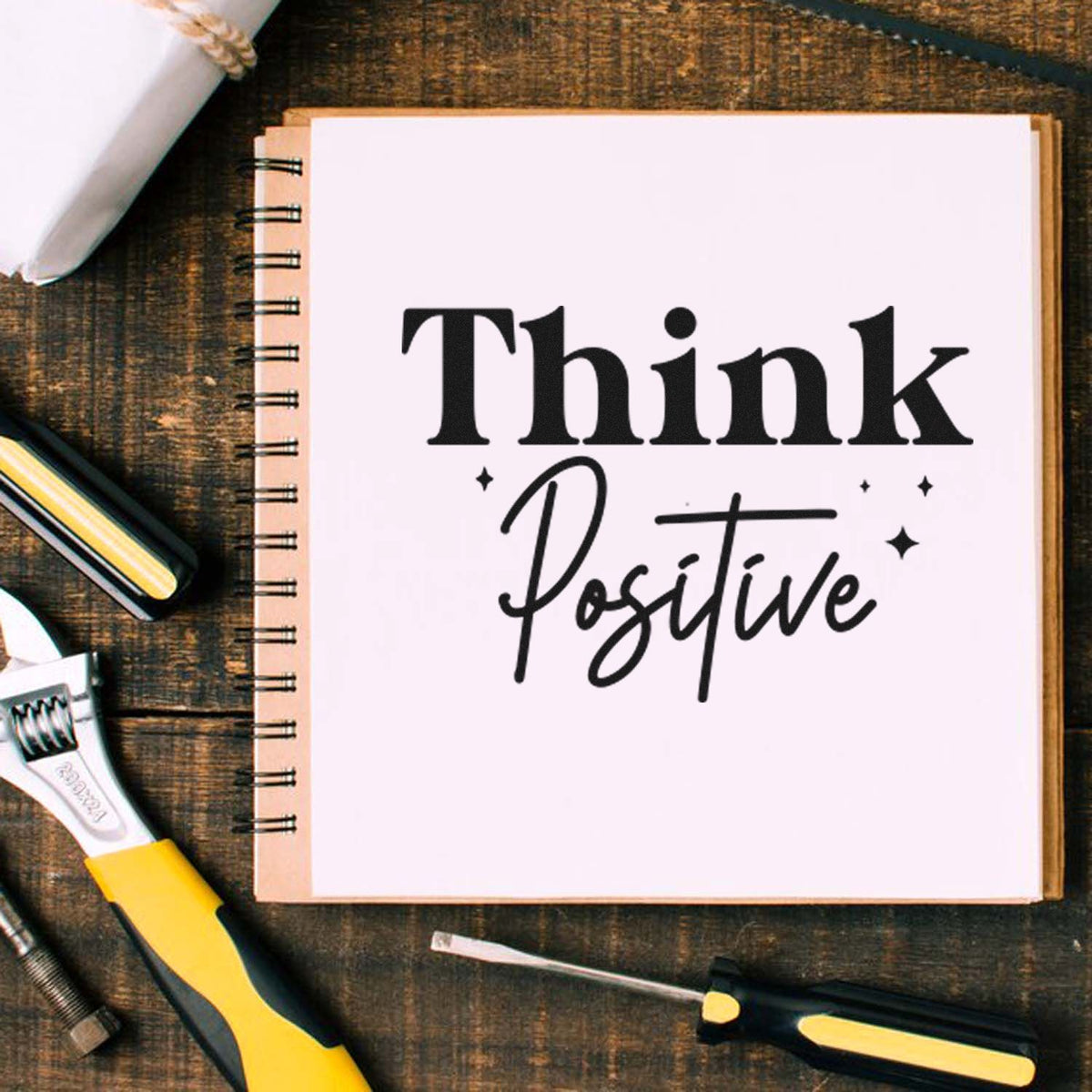 Custom stamp and seal set with black Think Positive lettering on a white notebook page, on rustic wood with yellow-handled tools.
