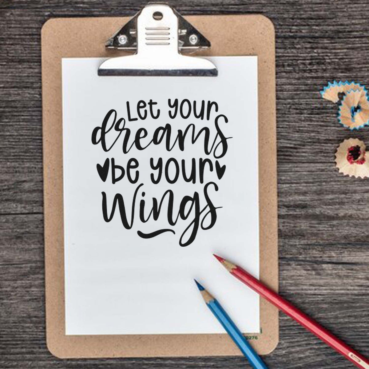 Brown clipboard with white paper reading Let your dreams be your wings in black script, plus red and blue pencils and decorative seal stamps.