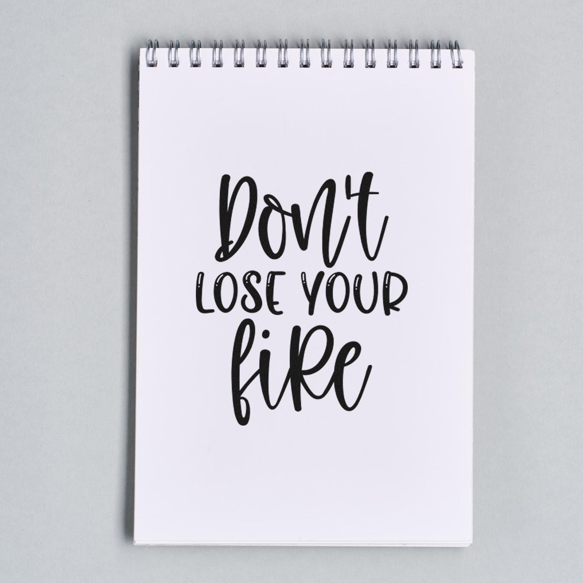 Black stamp-style motivational seal reading Dont lose your fire on a white spiral notepad.