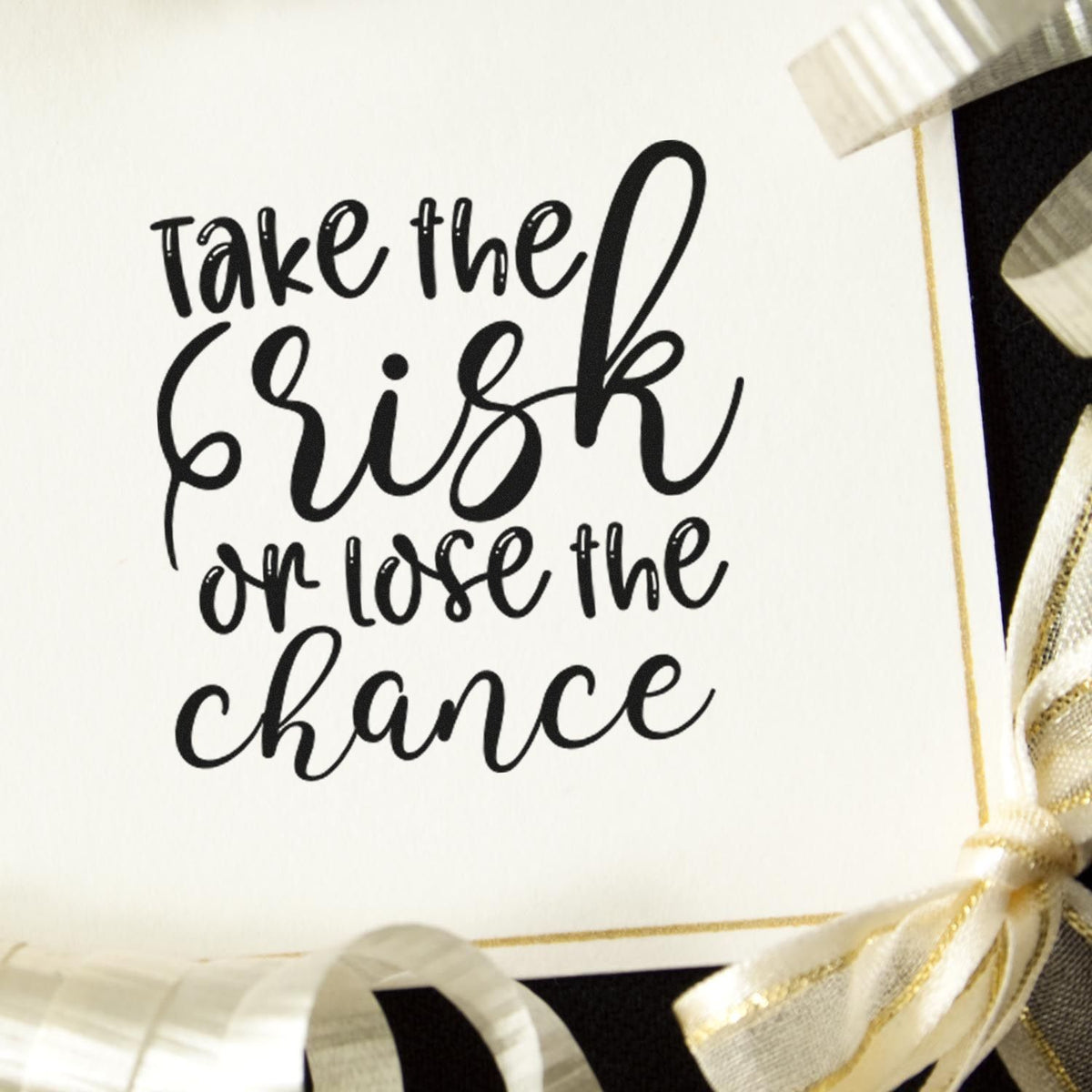 Black script quote stamp on white card with cream and gold ribbon seals: Take the risk or lose the chance.
