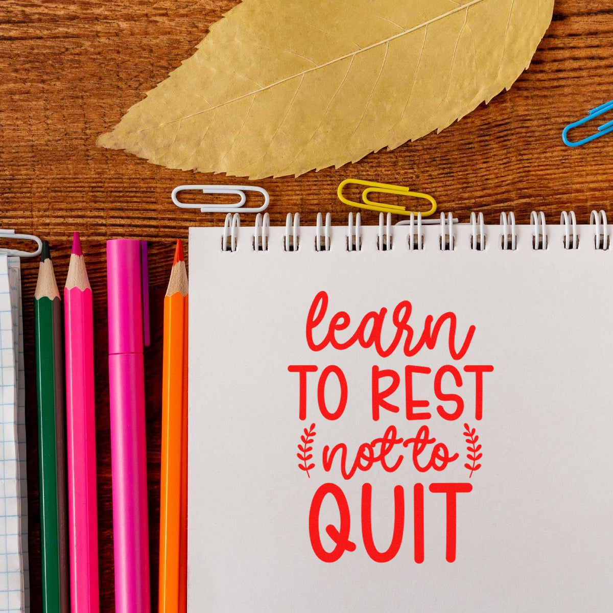 Red motivational stamp/seal on white spiral notebook, surrounded by pink, orange, and green pencils, paper clips, and a dried leaf.