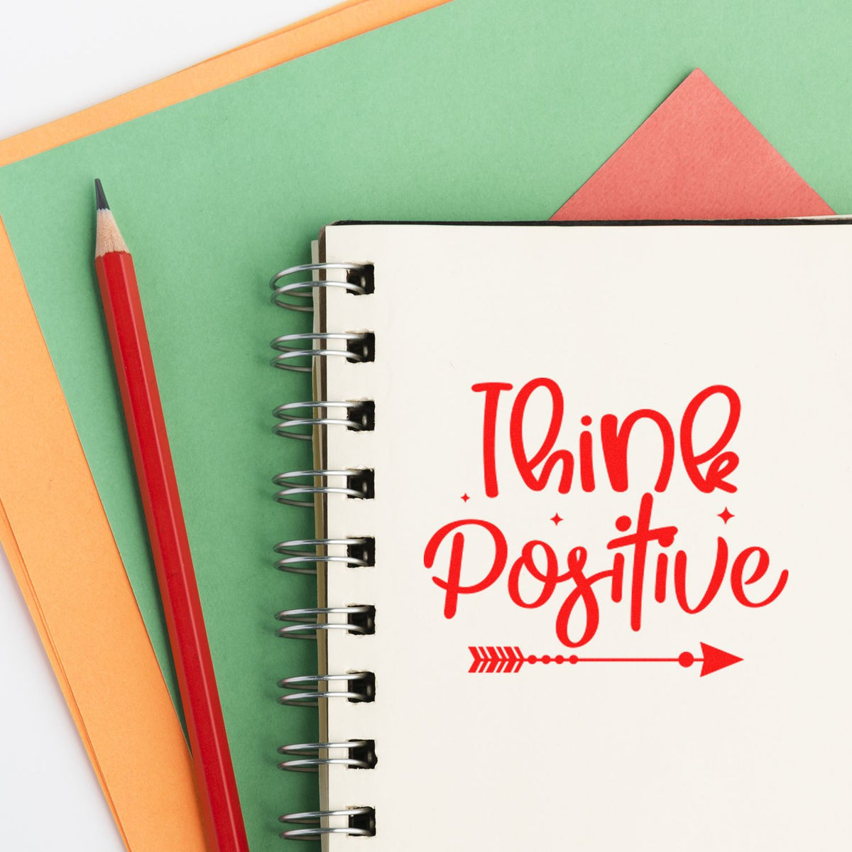 Red Think Positive stamp and seal design on a spiral notebook, with a red pencil and green, orange, and coral paper background.