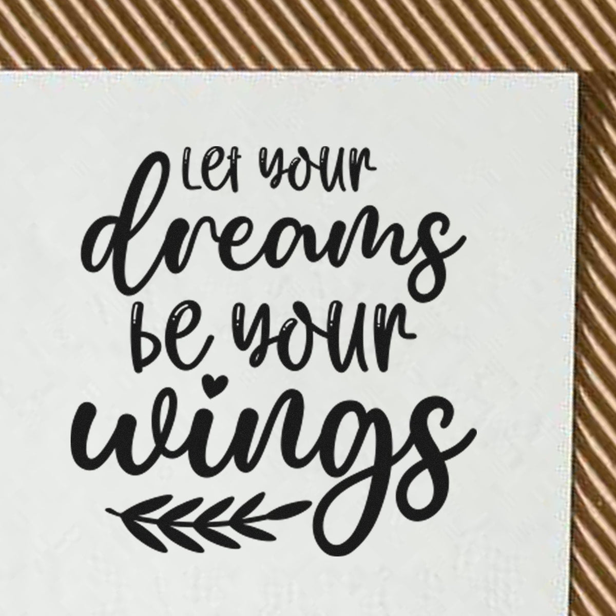Black motivational stamp seal reading let your dreams be your wings on white, with leaf accent, on brown striped background.