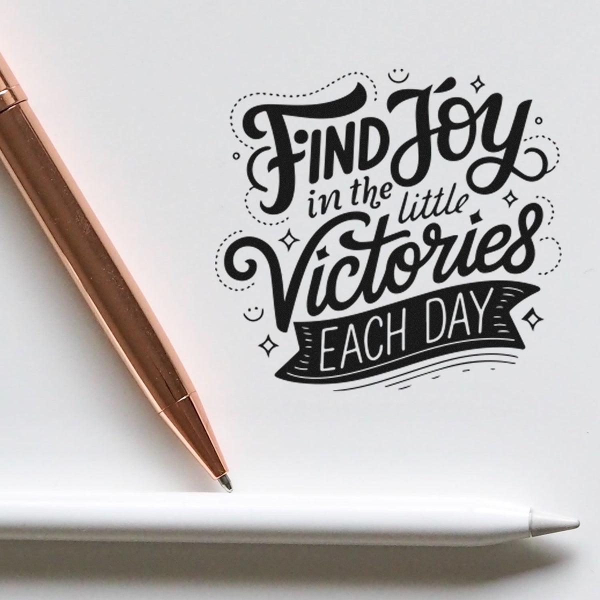 Black decorative stamp/seal reading Find Joy in the Little Victories Each Day on white paper, with copper pen and white stylus.