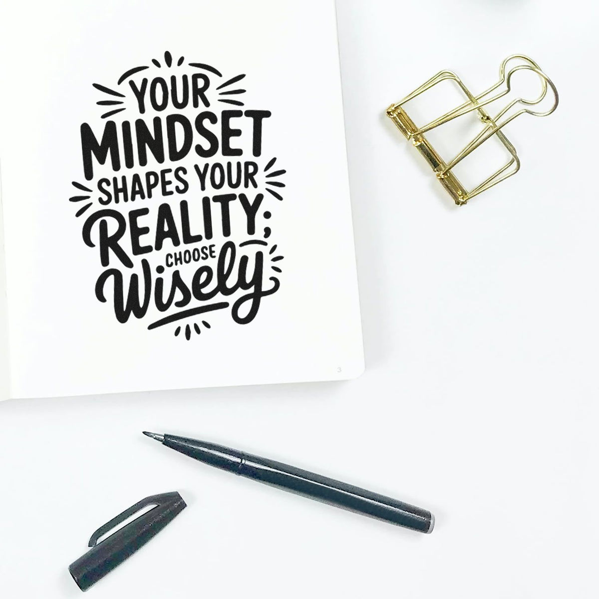 Black quote stamp and seal on white paper reads Your mindset shapes your reality; choose wisely, with gold binder clip and black pen.