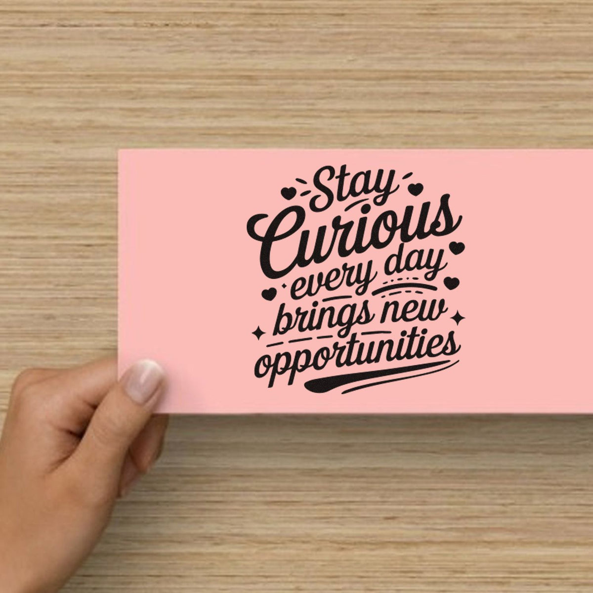 Pink decorative stamp seal card with black Stay Curious lettering, held by hand on a wood tabletop.