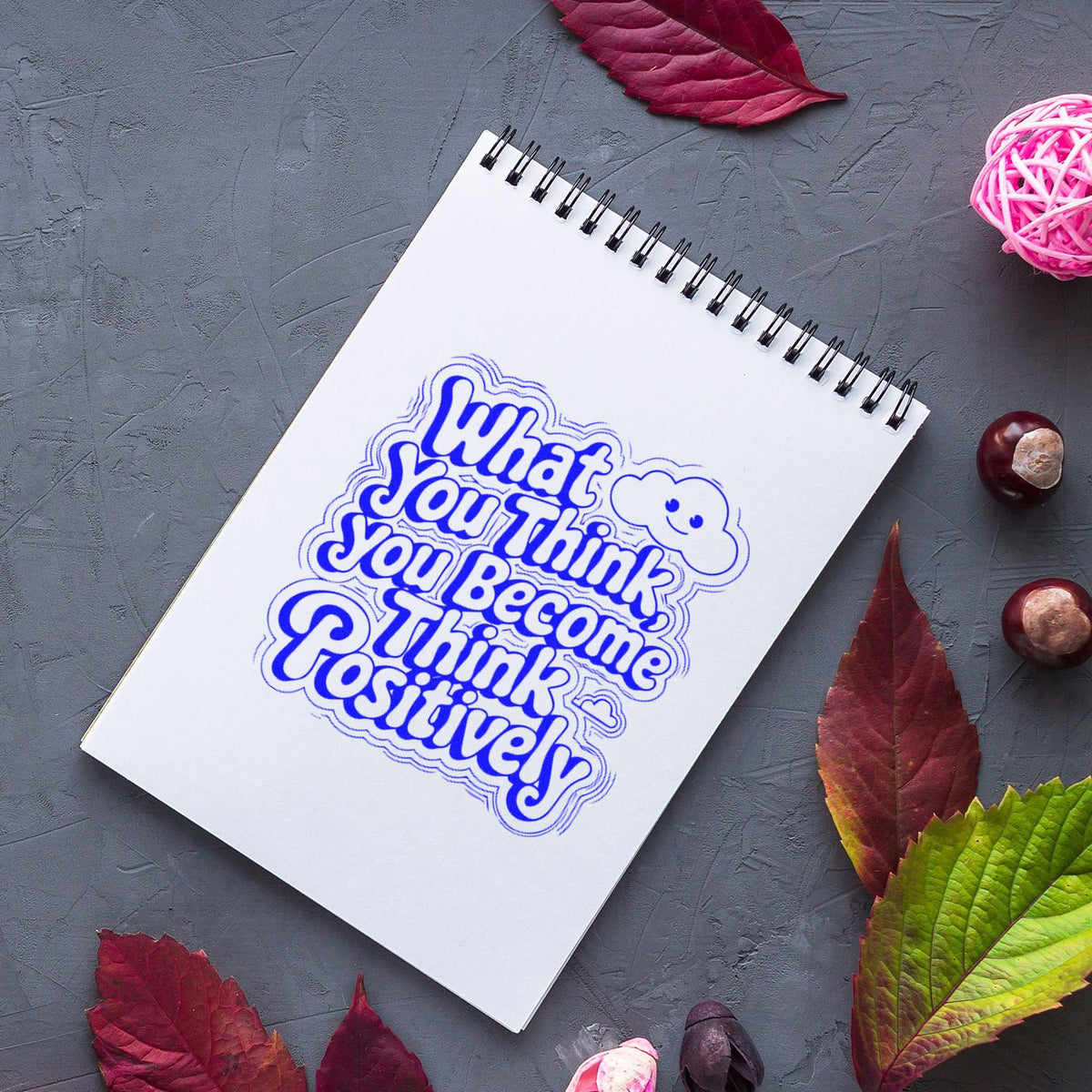 Blue custom rubber stamp seal on white spiral notebook, with What You Think You Become You Think Positively and autumn leaves.