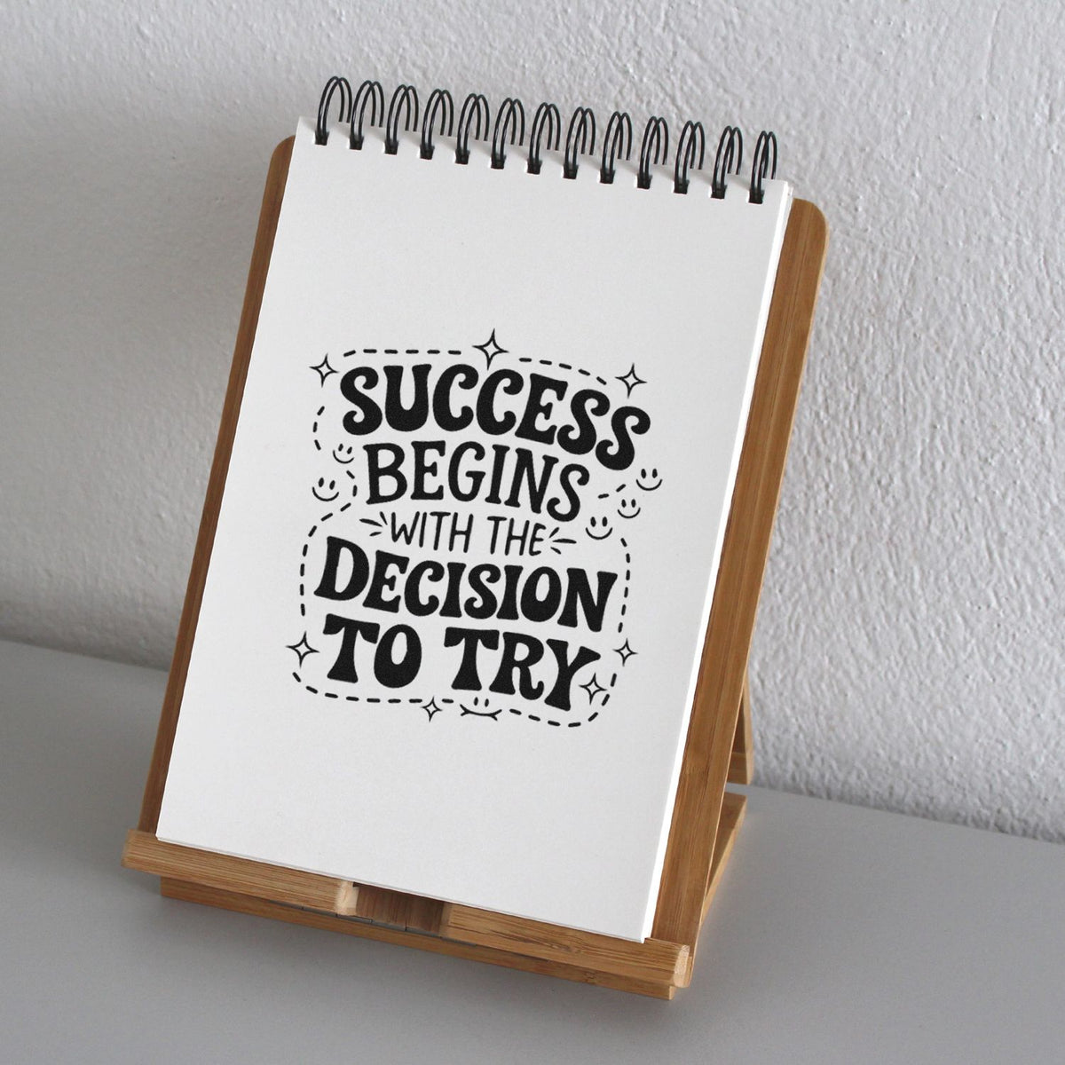 Black motivational stamp impression on white spiral notepad in wooden stand, reading Success begins with the decision to try.