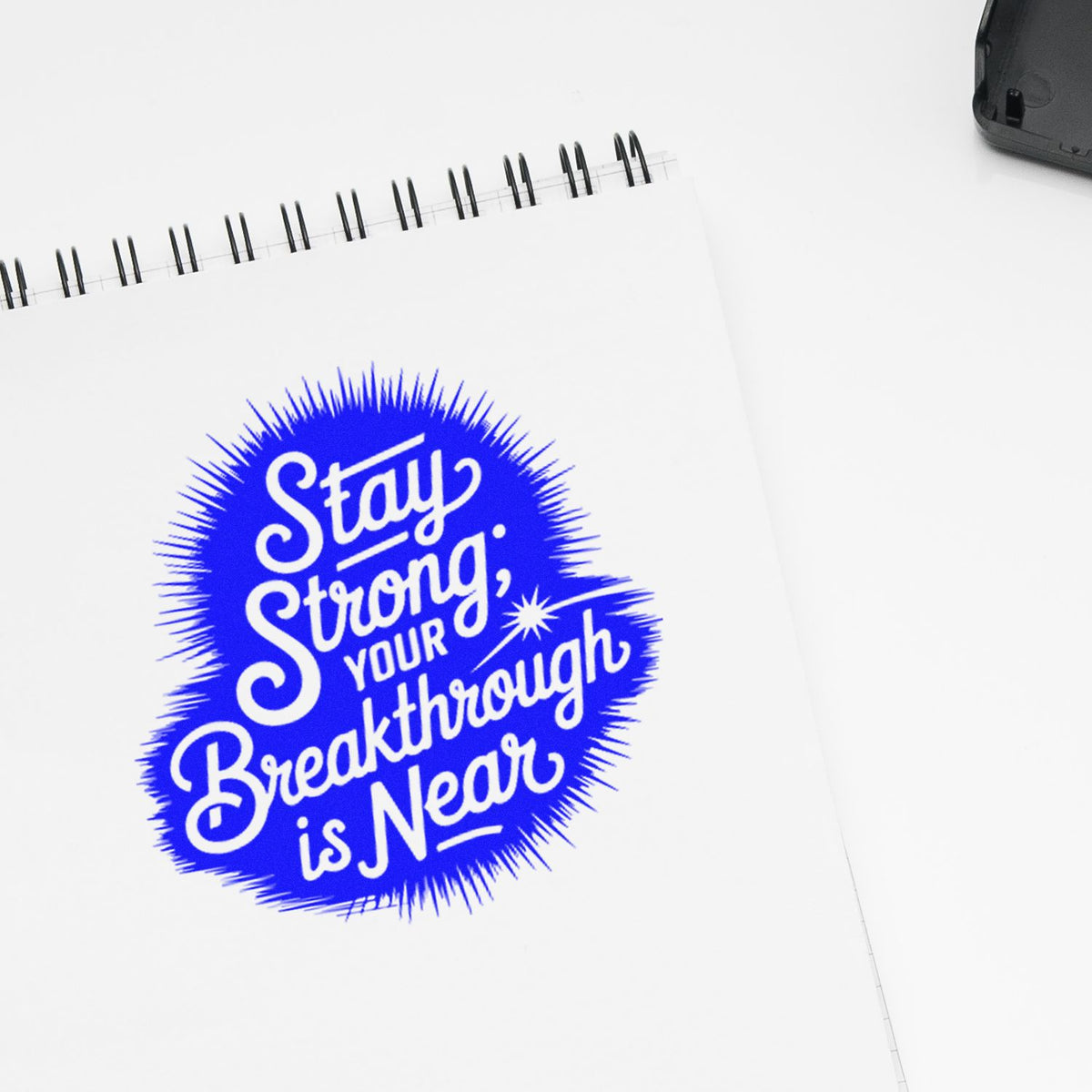 Blue rubber stamp seal with white lettering reading Stay Strong, Your Breakthrough is Near on a white spiral notebook.