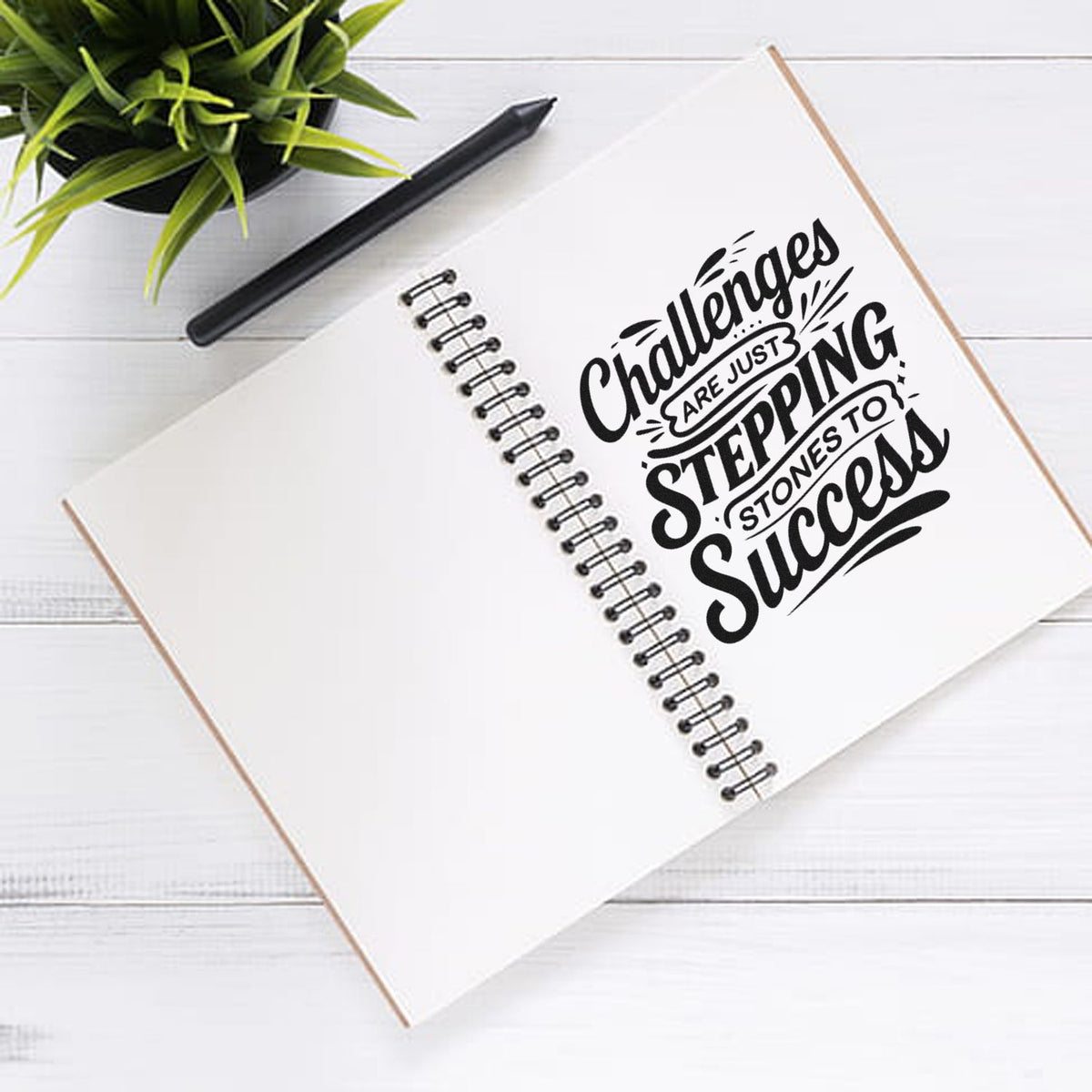 Black custom stamp/seal design on spiral notebook reading Challenges are just stepping stones to success, with pen and green plant.