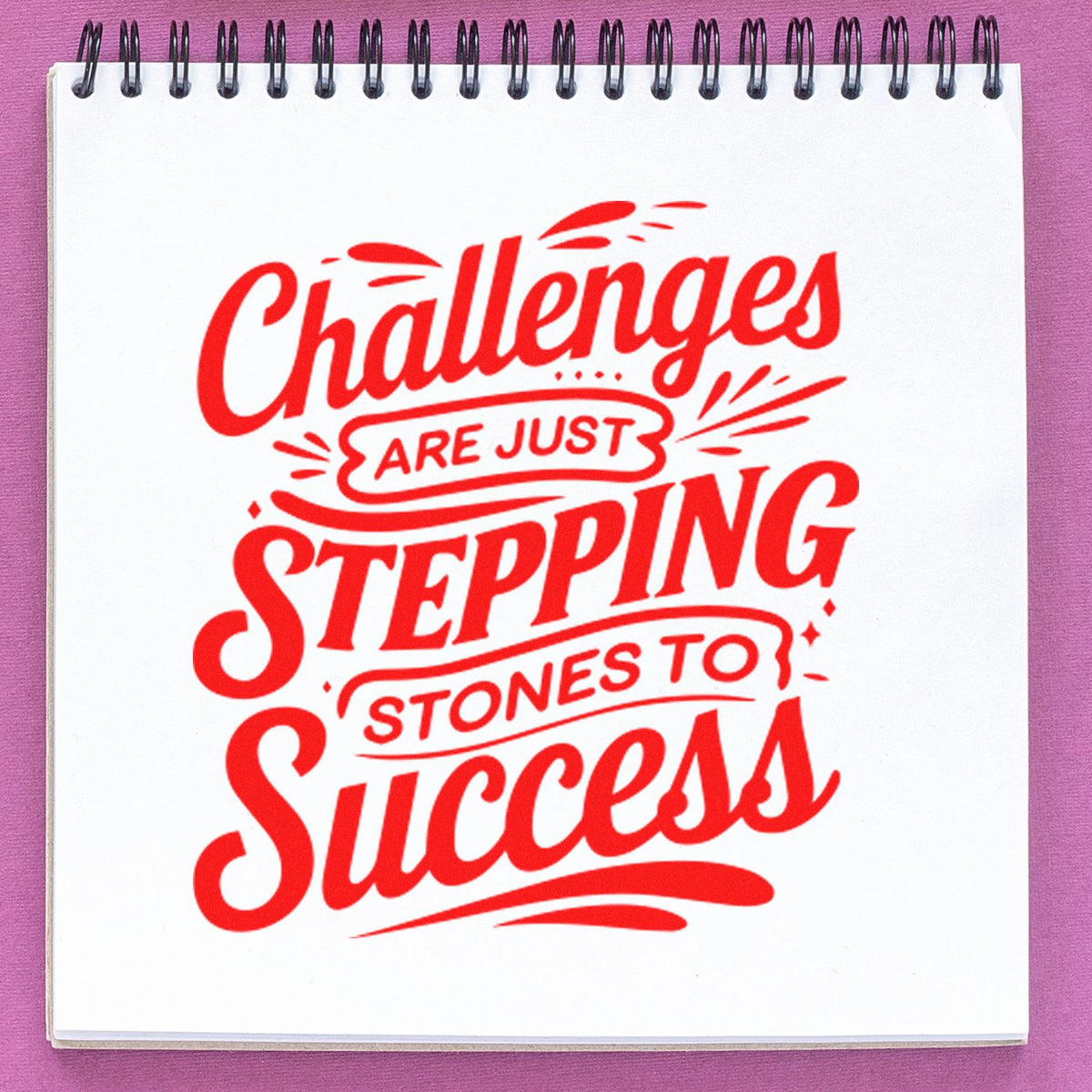 Red rubber stamp design on white spiral notebook reading Challenges are just stepping stones to success, on pink background.